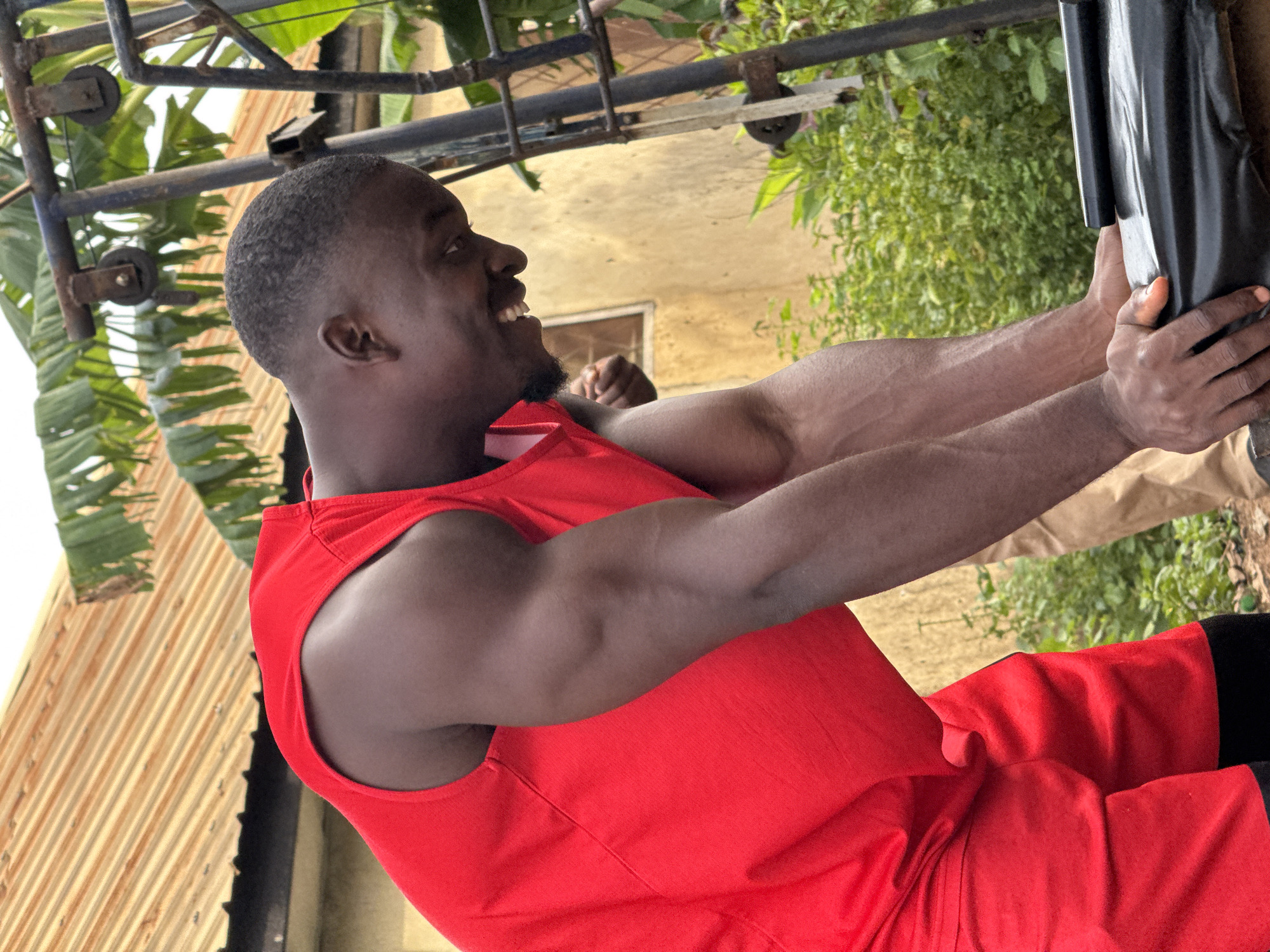The image shows a muscular Black man in a red athletic top and shorts, bending over and pushing down on a black object, possibly a tarp or cover. He is smiling broadly. The setting appears to be outdoors, with a corrugated metal roof and green foliage visible in the background. A metal structure with pulleys is also present. The man's pose and facial expression suggest he is engaged in some form of physical exertion or activity. The scene is likely located in the city of Akwanga, Nigeria.