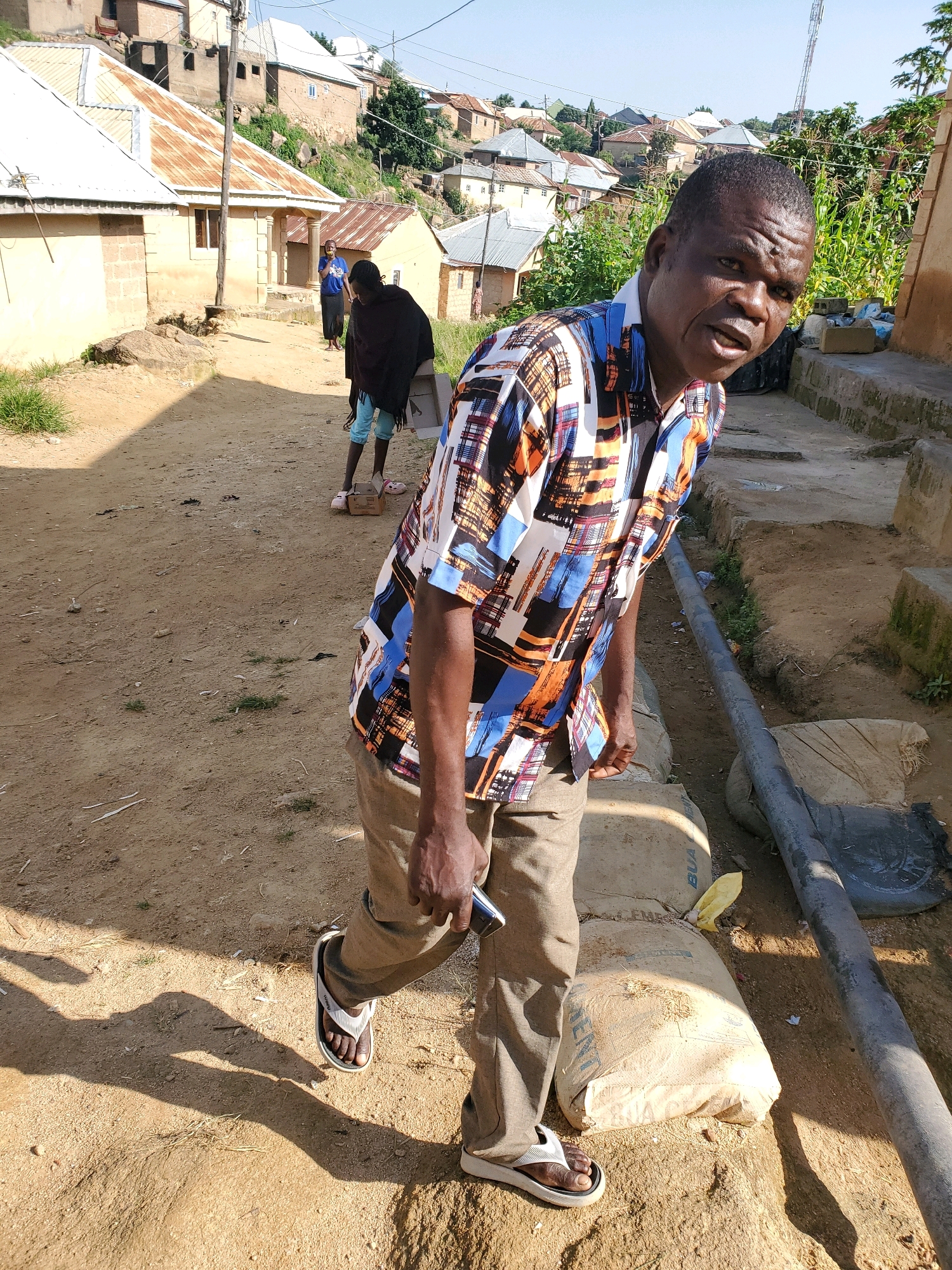 The image depicts an outdoor scene in a residential area of Jos, Nigeria, under a clear sky. In the foreground, a man wearing a colorful patterned shirt and khaki pants is standing and looking towards the camera. He has dark skin and short, dark hair. His left hand is holding a black mobile phone. He is wearing white and grey flip-flops. Behind him, a dirt path slopes upwards, with several buildings on either side. The buildings have corrugated metal roofs and some appear to be under construction.
Further in the background, two other individuals are visible. One, dressed in a dark garment and blue shorts, is standing near a building. Another person, wearing a blue shirt and dark skirt or pants, is also standing near a building.
The environment is characterized by a dusty, unpaved ground. Bags of what appear to be cement are stacked on the right side of the image, next to a large grey pipe. Vegetation, including some corn stalks, is visible in the background. The overall impression is of a community in a developing urban area.