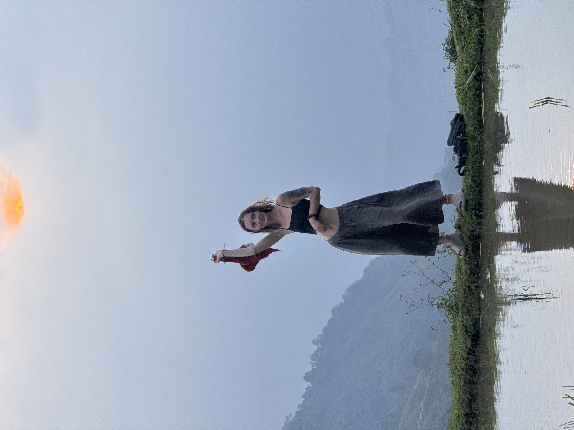 The image, when viewed upright, depicts a serene outdoor scene featuring a woman standing on a narrow grassy bank beside a calm body of water. In the distance, layers of mountains rise, partially obscured by a soft haze. The sky overhead is a clear, light blue.
The primary subject is a woman with light skin and dark hair, wearing a black crop top and dark, loose-fitting pants. She has a visible tattoo on her left arm and is looking towards the viewer with a relaxed expression, holding a red piece of fabric in her left hand. Her right hand rests on her hip.
To her right, on the grassy bank, a black backpack is visible. The water surface, likely a rice paddy or lake, perfectly reflects the sky and a warm, orange-yellow light source visible on the left side of the frame, suggesting either a sunrise or sunset. The light also highlights elements on the water's surface, enhancing the reflective quality. The vegetation on the bank is green and somewhat wild. The overall atmosphere is tranquil. The mountainous and watery landscape aligns with the setting of Nậm Dịch Commune, Vietnam.