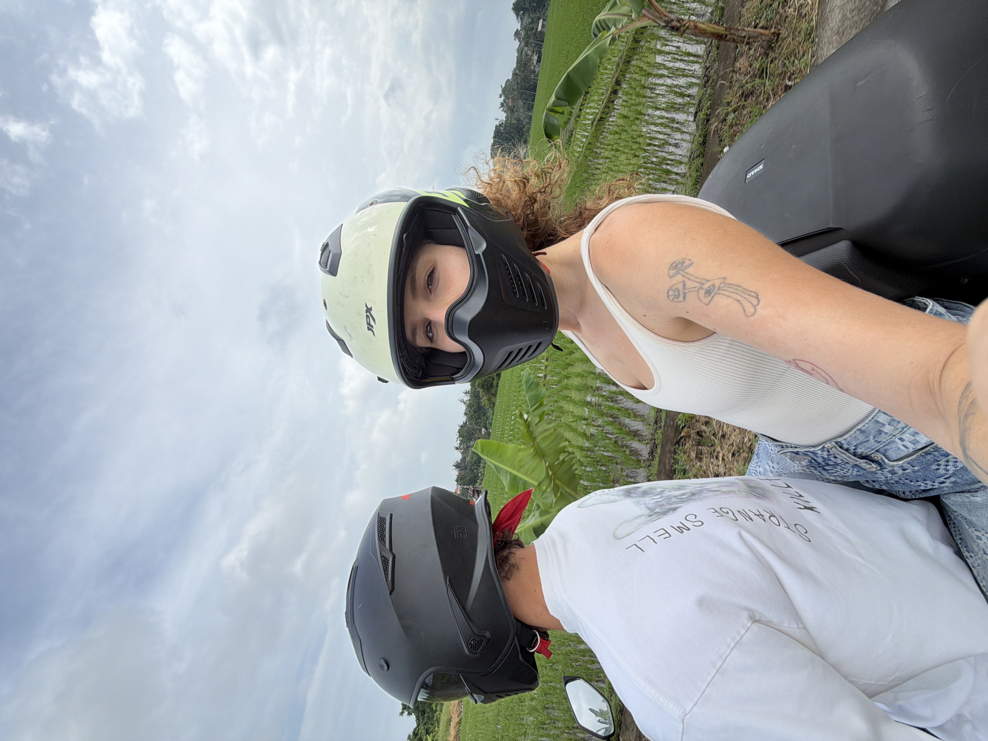 Two individuals are seated on a motorcycle, with the foreground occupied by the back of a person wearing a black helmet and a white t-shirt with text. To the right, a person wearing a light-colored helmet and a white tank top faces the camera, looking towards the viewer. Both individuals are wearing helmets, suggesting an outdoor activity. The background reveals a lush, green rice paddy field, characteristic of rural Indonesian landscapes. A few banana trees are visible among the rice stalks, and the sky is overcast with clouds. The scene appears to be in North Kuta, Indonesia, given the presence of rice paddies and tropical vegetation. The overall impression is a casual, outdoor moment captured during a motorcycle ride through a scenic agricultural area.