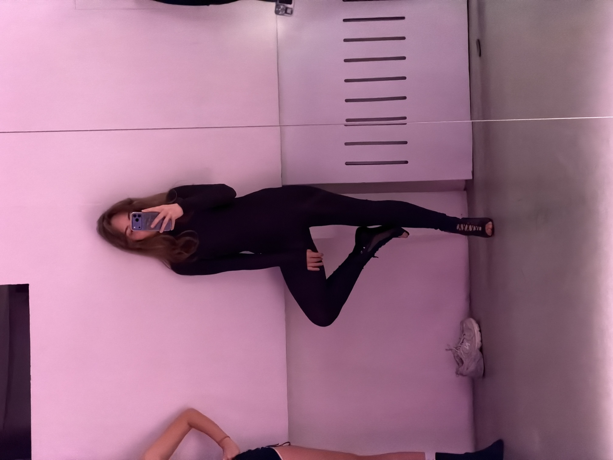 A woman in a black outfit is taking a selfie in a room with a mirrored wall. Below the mirror, another person is lying down wearing shorts. A pair of sneakers are on the floor. The environment is predominantly pink and gray. There is no information to specify that it is in the city of Moscow, Russia.