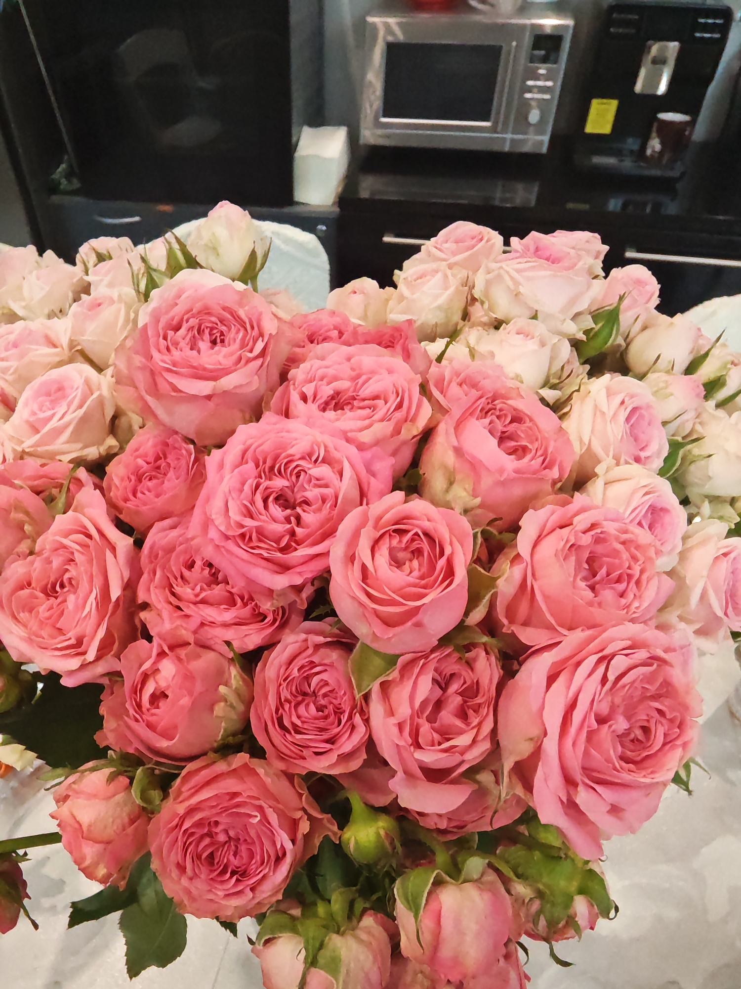 A vibrant bouquet of pink roses dominates the foreground, filling most of the frame. The roses are in various stages of bloom, with many fully open, showcasing their dense, layered petals, while others are still tight buds. The predominant color is a rich, medium pink, with some roses exhibiting lighter pink or almost white edges, especially among the buds. Green leaves and stems are visible interspersed throughout the bouquet, adding a touch of freshness.
In the blurred background, typical kitchen elements are discernible. On the left, a silver-toned microwave oven with a dark screen and buttons is visible. To its right, a dark, glossy coffee machine with silver accents and a yellow, unreadable label is present. Both appliances rest on a dark, possibly black, glossy countertop, which reflects some light. A white, rectangular object, possibly a tissue box, is also on the counter. The overall setting appears to be an indoor space, likely a kitchen, with artificial lighting. There are no people visible, and no specific activity is taking place other than the static display of the flowers. No legible text is visible on any objects. The scene conveys a sense of domesticity and beauty. The location context for this media file is Rybolovo, Russia.