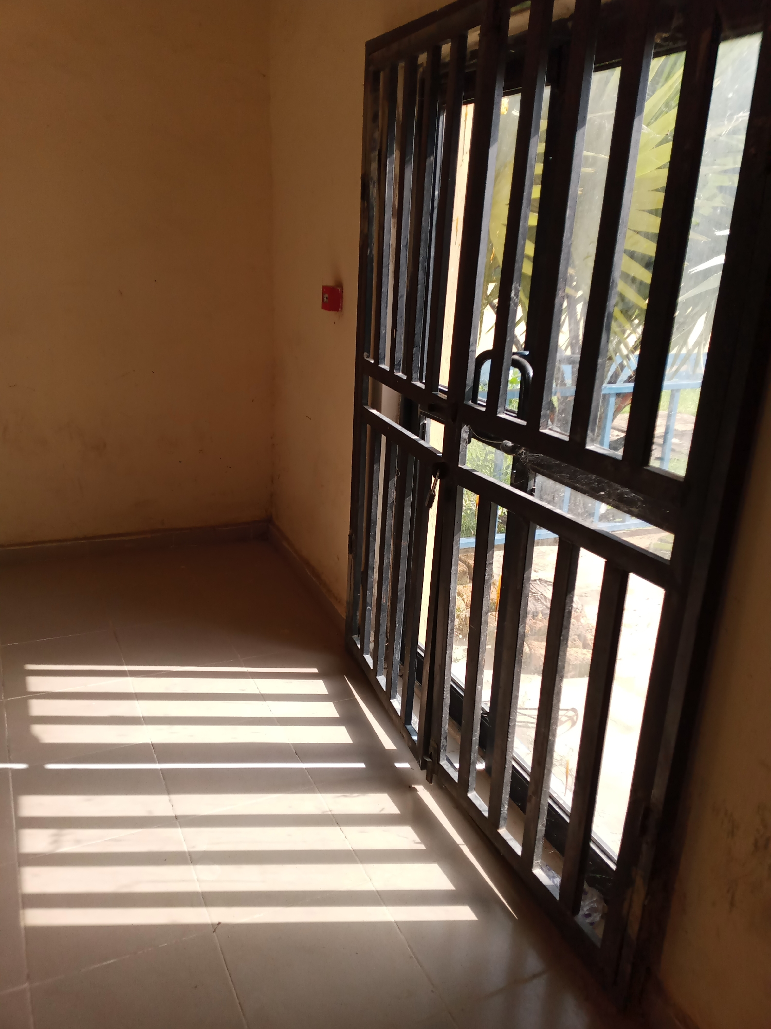 The image depicts an interior view of a room with beige walls and tiled flooring. A metal security gate with vertical bars covers a glass door, allowing sunlight to cast distinct parallel shadows across the floor. To the left of the gate, mounted on the wall, is a red fire alarm. The scene appears to be an unoccupied interior space. The view through the gate reveals a hint of outdoor greenery, suggesting it might be a building in Jalingo, Nigeria, given the lighting and common security features.
