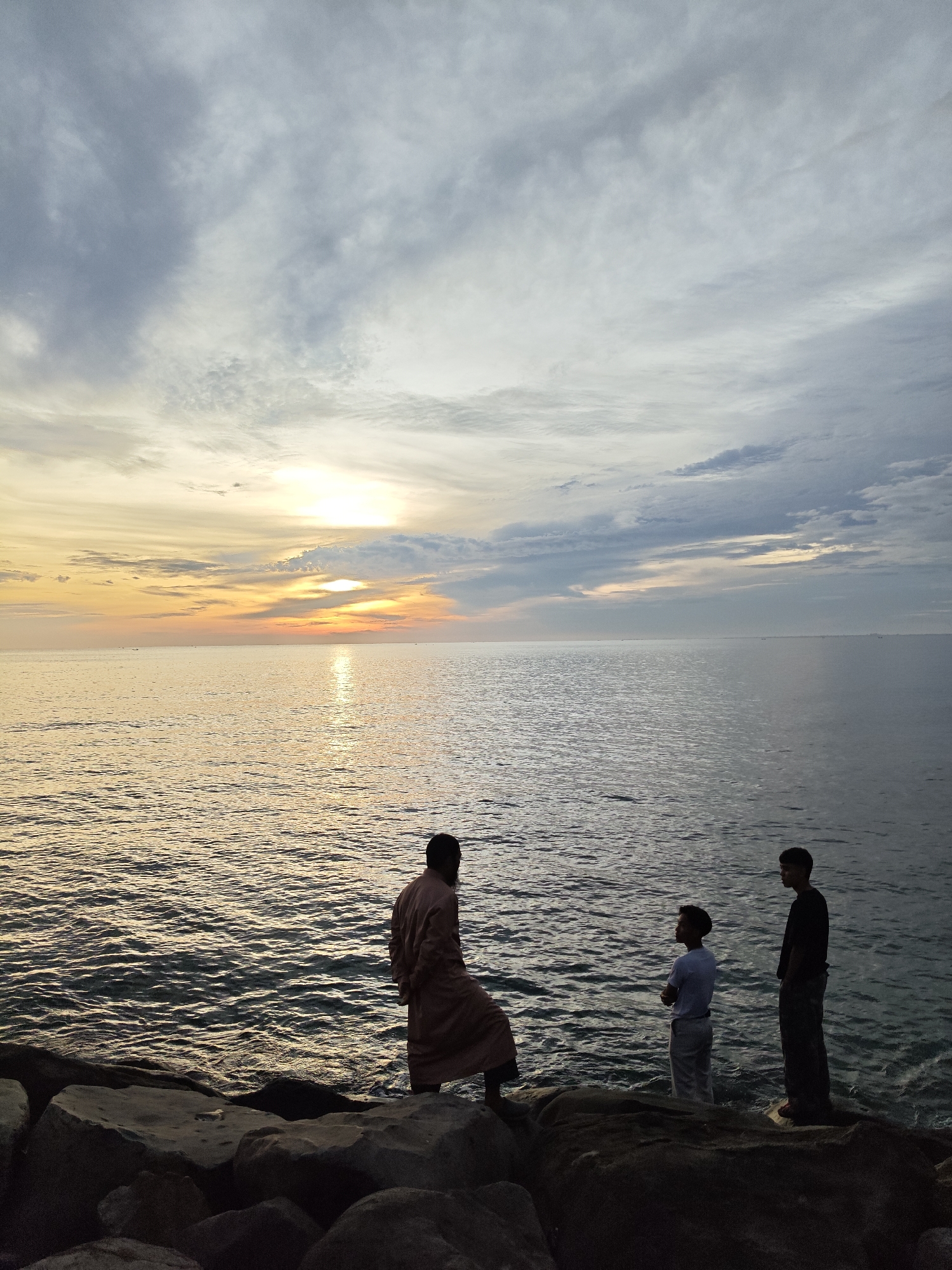 An image captures a sunset scene on the coast of Lhokseumawe, Indonesia, with three men standing on a rocky shore. The sun, positioned near the horizon on the left side of the frame, casts a bright, elongated reflection across the calm ocean water, illuminating it with golden hues. The sky above features scattered clouds, displaying a gradient of warm oranges and yellows near the sun, transitioning to light grays and whites towards the top and right.
In the foreground, a dark, uneven rock formation extends along the bottom of the image. Three adult males are standing on these rocks, facing the ocean. The leftmost individual, positioned on a large rock, wears a long, reddish-brown robe and is seen from the back, slightly turned towards the right. To his right, a second man stands closer to the water's edge, dressed in a white short-sleeved shirt and light-colored trousers, with his arms crossed as he looks out at the sea. The third man, located furthest to the right and also near the water on a rock, wears a dark long-sleeved shirt and dark trousers, and is also facing the ocean. All three figures are partially silhouetted against the bright background of the sunset. Several small, indistinct vessels are visible on the distant horizon line across the ocean.
