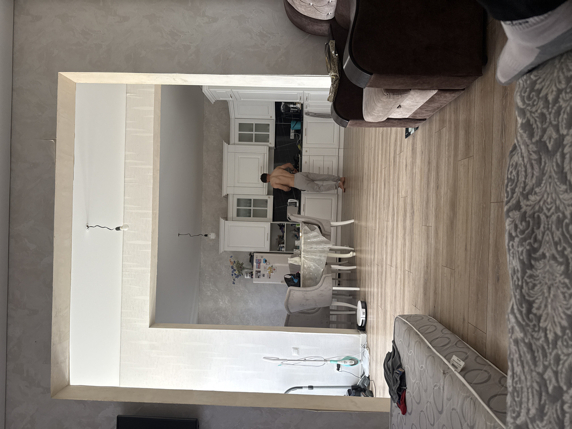 The image depicts an interior scene, likely a modern apartment or house in Yasnaya Polyana, Russia. The main subject is a man with his back to the camera, wearing grey pants and no shirt. He appears to be standing in a kitchen area, facing white cabinetry and a dark countertop. The kitchen is well-lit and appears to be part of a larger open-plan living space.
To the right of the kitchen, a dining area is visible with a table and chairs. The table is covered with a clear plastic tablecloth, and there are several chairs around it. Further to the right, a portion of a sofa and a person's legs, clad in grey and white socks, are visible, suggesting a living room area adjacent to the dining and kitchen space.
In the foreground, a mattress is partially visible, draped with a grey and white patterned fabric. Adjacent to the mattress, various cleaning appliances are present, including a vacuum cleaner and a steam mop.
The scene appears to be indoors during daylight hours, with artificial lighting contributing to the overall brightness. The décor is modern, with light-colored walls and wooden flooring. The overall impression is one of a lived-in space, possibly in the process of being cleaned or organized. There is no discernible text visible in the image.