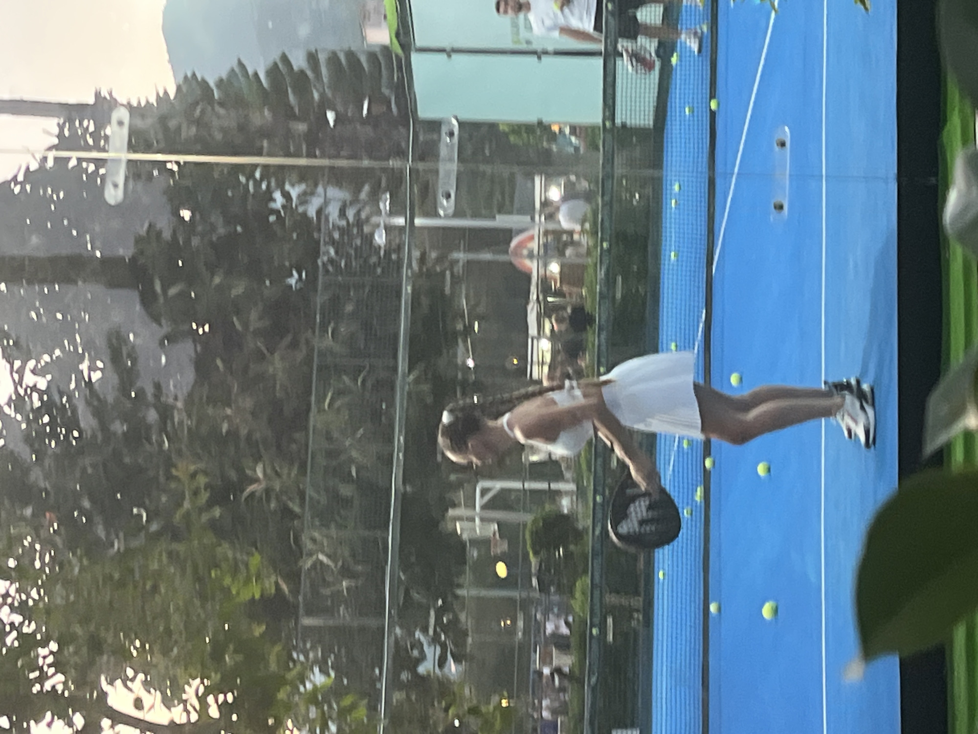 A medium shot, taken in an outdoor setting, reveals a woman playing padel tennis on a blue court with a glass wall. The woman, dressed in white, is in mid-swing with a black paddle. The padel tennis court is lined with yellow tennis balls. Through the glass wall, there's a view of a city scene. This is the city of Alanya in Turkey, characterized by trees, buildings, and a mountain range in the background. Other individuals are visible in the background as well.