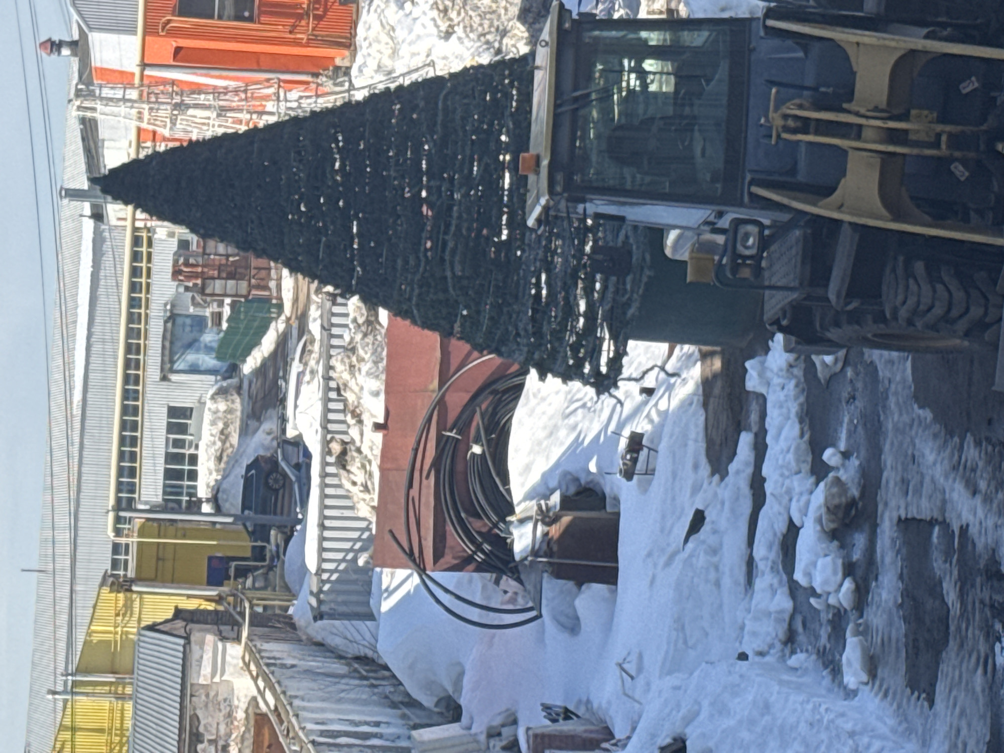 The image displays an outdoor industrial or storage area in Chaikovsky, Russia, under conditions indicating winter or early spring, with significant snow, slush, and ice coverage. In the immediate foreground, positioned to the right, is the front section of a yellow and dark grey front-end loader, featuring a yellow bucket assembly and an orange beacon light on its cab roof. The ground beneath consists of dark asphalt, melting snow, and standing water.
A tall, conical, dark green artificial Christmas tree, constructed from layered artificial foliage, stands centrally in the midground. To the left of the tree, a substantial snow-covered embankment is present, behind which a large, rectangular, dark red metal structure is visible. Several coiled black pipes or cables are arranged against this red structure and on an adjacent snowdrift. Further left, additional snow covers miscellaneous debris and a smaller, rust-colored metal container. A horizontal corrugated metal fence acts as a boundary across the midground.
The background comprises an industrial complex featuring multiple buildings. A prominent yellow corrugated metal building is situated on the left, next to a larger building with a grey corrugated roof. An orange corrugated metal building is visible on the right. Various metal pipes, structural elements, and a ladder are scattered throughout the background. A dark-colored SUV is parked near the yellow building, partially obscured by snow. The sky overhead is clear and light blue. No individuals are visible in the scene.