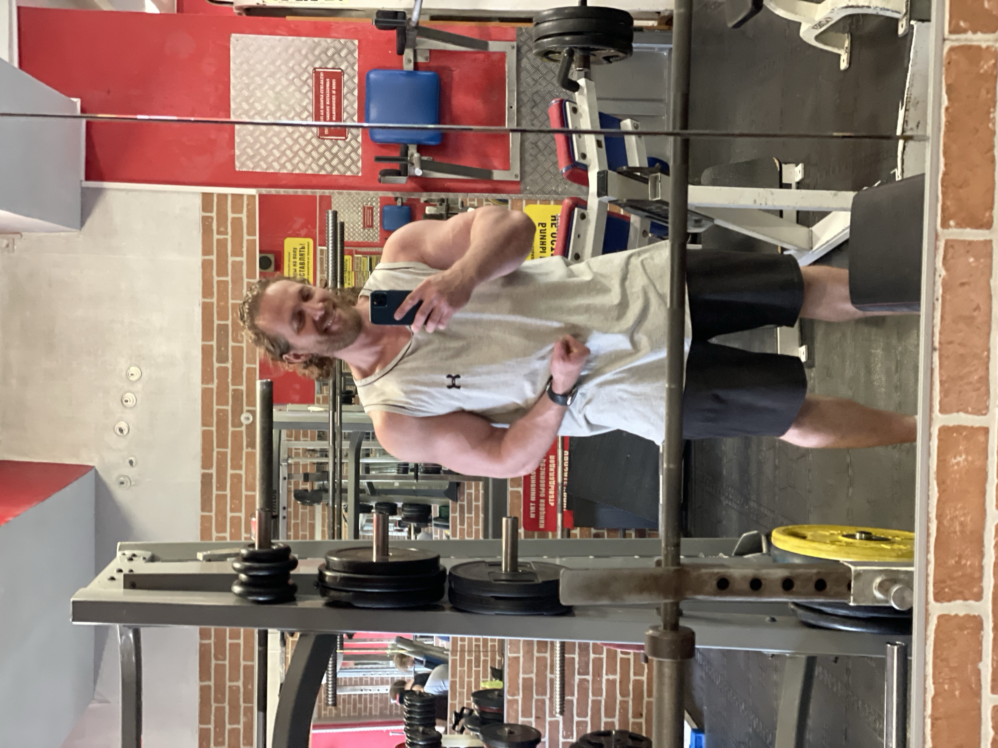 The image shows a man taking a selfie in a gym. He is wearing a grey tank top and black shorts, and he has curly blonde hair. He is smiling and flexing his bicep. The gym appears to be in a brick building, with red and white walls and various weightlifting equipment visible in the background. There are also other people in the gym, but they are out of focus. The man is holding a smartphone in his right hand, and he is looking at his reflection in a mirror. The gym is located in Saint Petersburg, Russia.