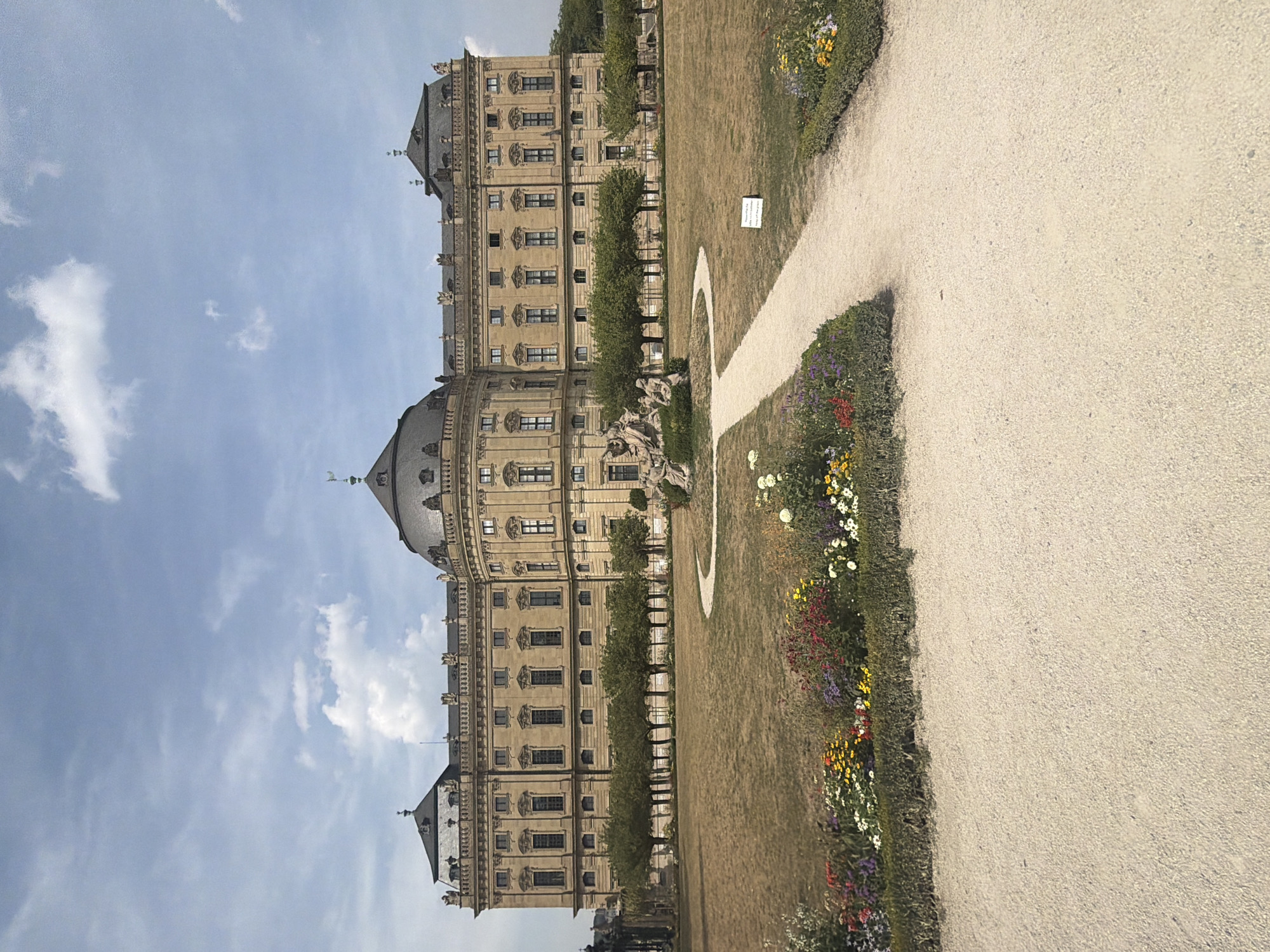 The image shows the Würzburg Residence, a UNESCO World Heritage site located in Würzburg, Germany. The grand palace, with its symmetrical Baroque architecture, is the central focus. It features a large, light-colored facade with numerous windows and ornate detailing. The palace is flanked by dark, multi-gabled roofs and topped with a prominent dome. In front of the palace, a meticulously maintained garden stretches out. A pathway, made of light-colored gravel, curves through the lawn. The lawn itself appears dry and somewhat parched, suggesting a sunny day. To the left of the pathway, a hedge separates a vibrant flowerbed filled with a variety of colorful blooms. In the background, a row of evenly spaced trees forms a line between the palace and the lawn. A statue is visible in the middle ground, partially obscured by trees and the curve of the path. The sky above is blue with scattered white clouds. The overall scene conveys a sense of historical grandeur and formal landscaping.