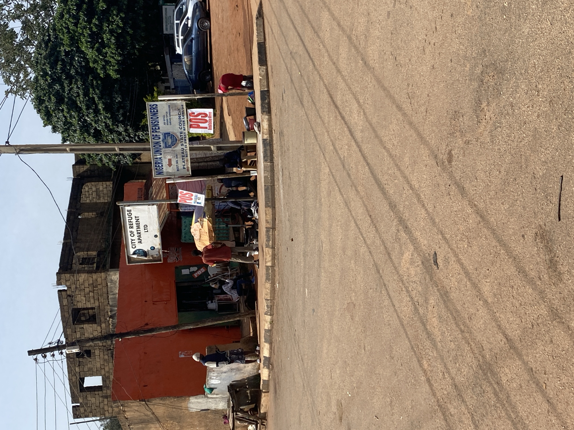 The image depicts a street scene in Gura Top, Nigeria. In the foreground, a paved road with tire marks and shadows from overhead wires is visible. A sidewalk with a curb separates the road from the buildings. Along the sidewalk, several people are gathered. On the left, a person in a blue jacket and white head covering stands near a wooden stall. Further down, another person in a red shirt stands. In the middle, several individuals are seated or standing near shop entrances. To the right, a person in a red garment is attending to some items on the ground. Two cars are parked on the side of the road. A large, leafy green tree is prominent in the background. Signs for "CITY OF REFUGE APARTMENT LTD" and "NIGERIA UNION OF PENSIONS" are visible on the buildings, along with "POS" signage indicating point-of-sale services. The buildings appear to be a mix of brick and painted structures, some appearing unfinished. The overall environment suggests a busy urban street with commercial activity.