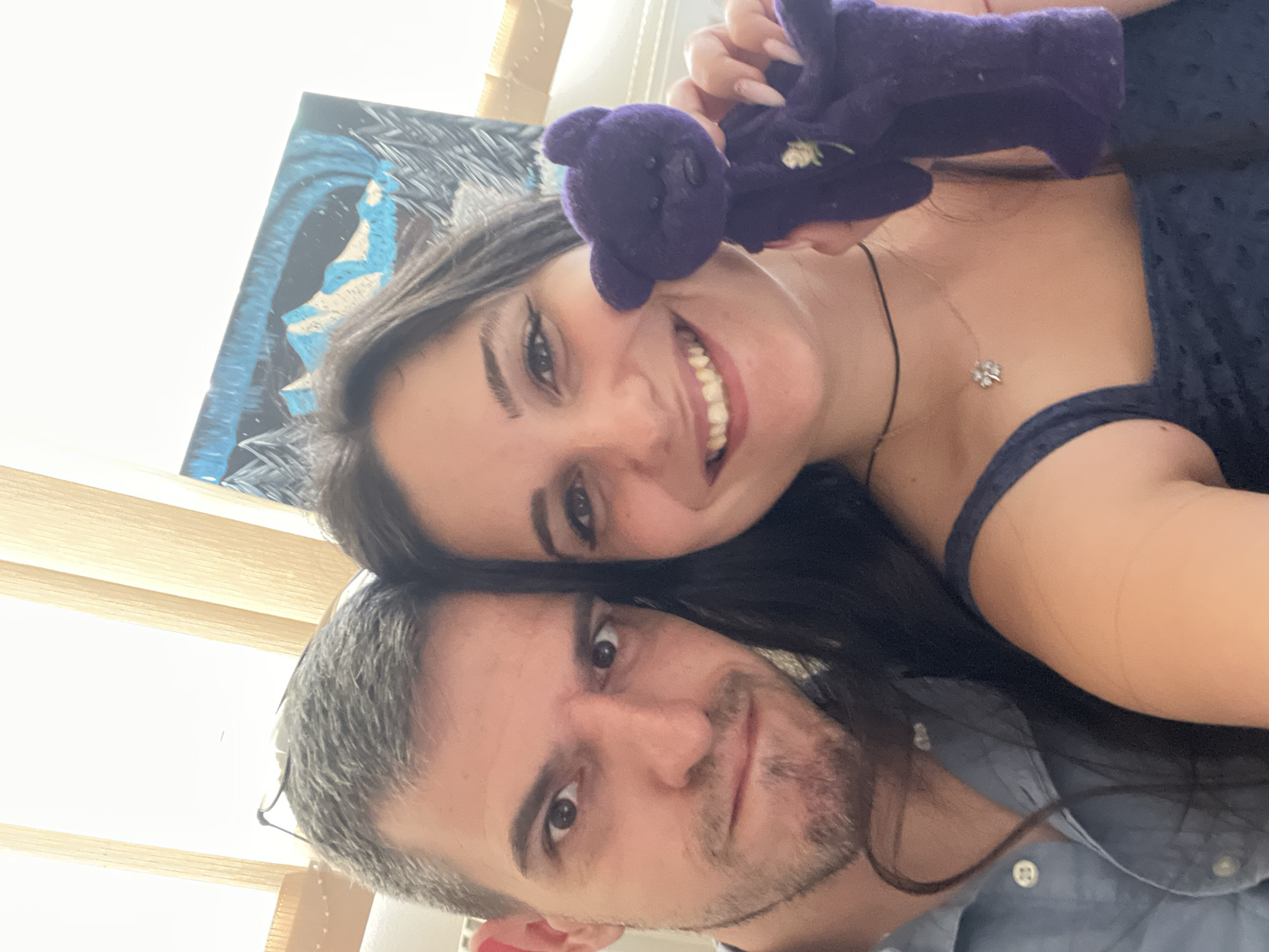 The image shows a close-up selfie of a man and a woman, likely a couple. The man, on the left, has short graying hair and is wearing a blue collared shirt. He is looking directly at the camera with a slight frown. The woman, on the right, has long dark hair and is smiling. She is holding a small purple teddy bear with a white flower on its chest. She is wearing a dark blue top and a thin black choker necklace with a silver pendant shaped like a four-leaf clover. Behind them, vertical blinds are visible, suggesting an indoor setting. A painting of a winter landscape with mountains and the aurora borealis is also in the background. The lighting is bright, indicating daytime. This scene is located in Sofia, Bulgaria.