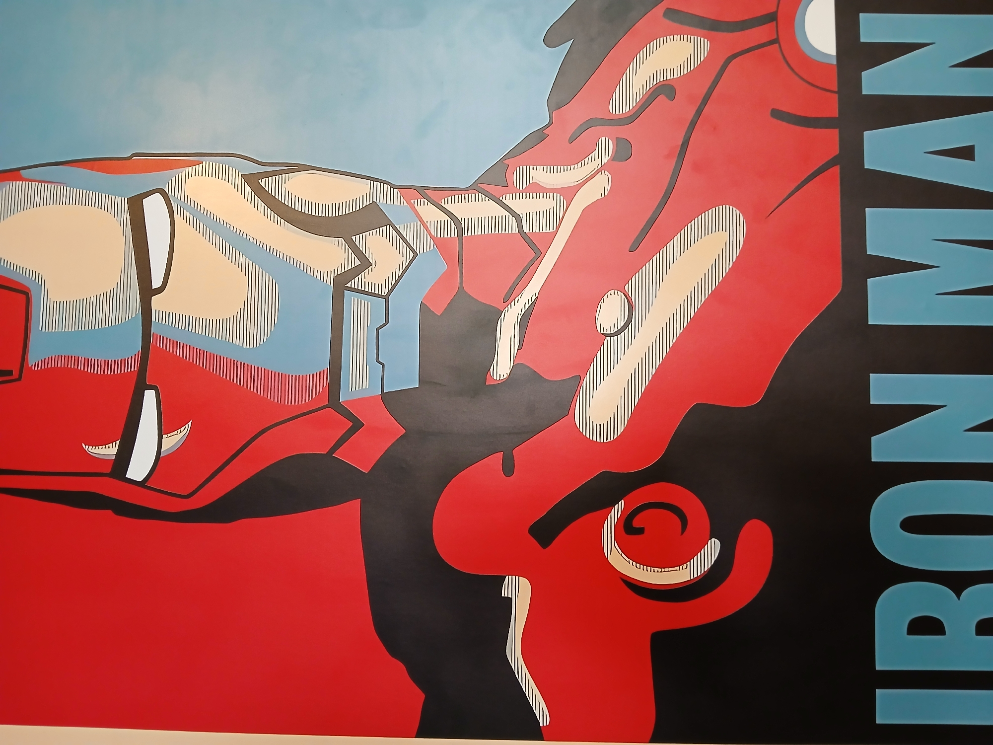 A mural of Iron Man, rendered in a stylized, graphic art style, fills most of the frame. The artwork uses bold, flat colors of red, black, beige, and blue, with black linework and striped fills. The right side features the blue text "IRON MAN" vertically against a black background. The environment appears to be an indoor wall. No people are visible. The location is unknown.