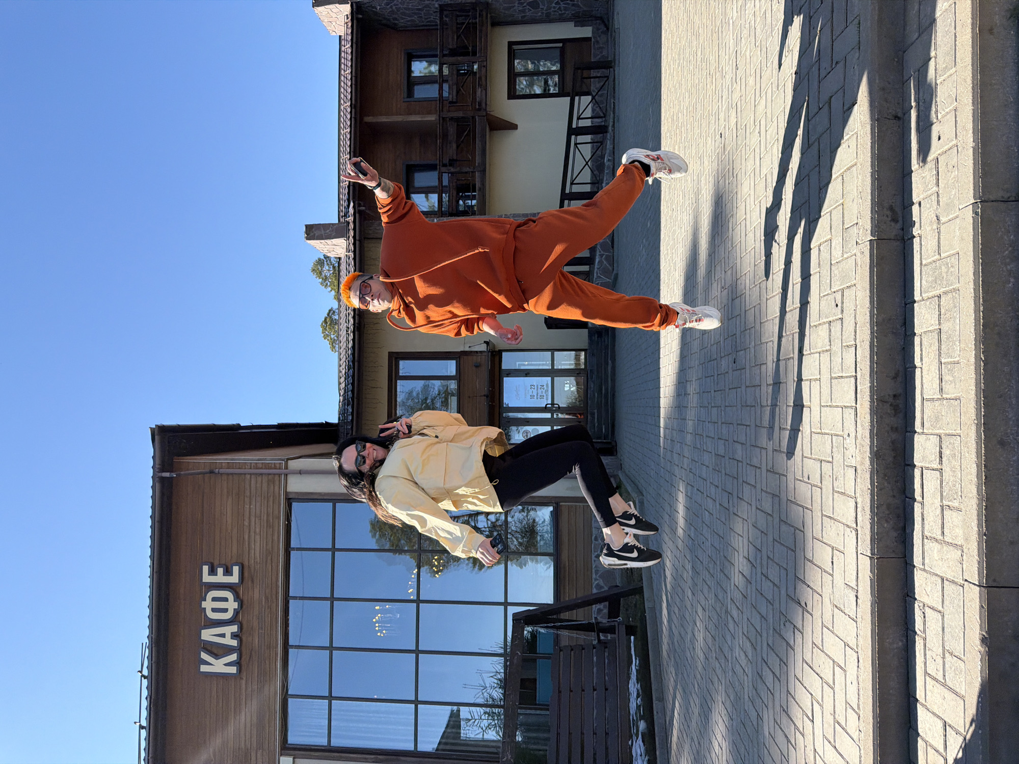 Two people are posing for a photo outside a building. One person, wearing an orange jumpsuit and glasses, is striking a dynamic pose with one leg extended. The other person, wearing a light-colored jacket and black leggings, is making a peace sign with their fingers. The building is labeled "КАФЕ" in Cyrillic letters. The image appears to have been rotated. The location is likely Votkinsk, Russia.