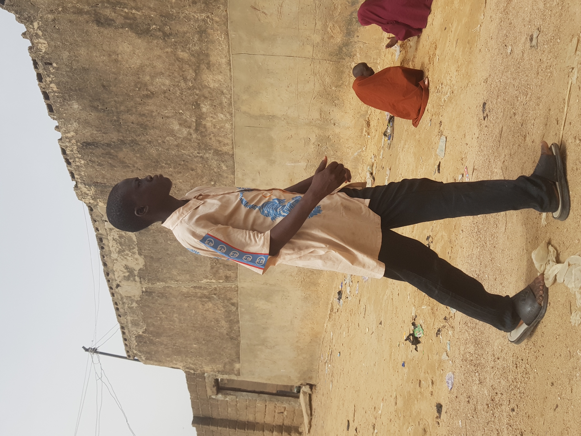 The image shows a young man standing in a dirt area next to a building with a weathered concrete exterior, possibly in Dutsinma B, Nigeria. He is wearing black pants, a light-colored patterned shirt, and dark sandals. He is looking up towards the building. Two other people are visible in the background, kneeling on the ground. There is trash scattered on the ground. Overhead wires are also visible.