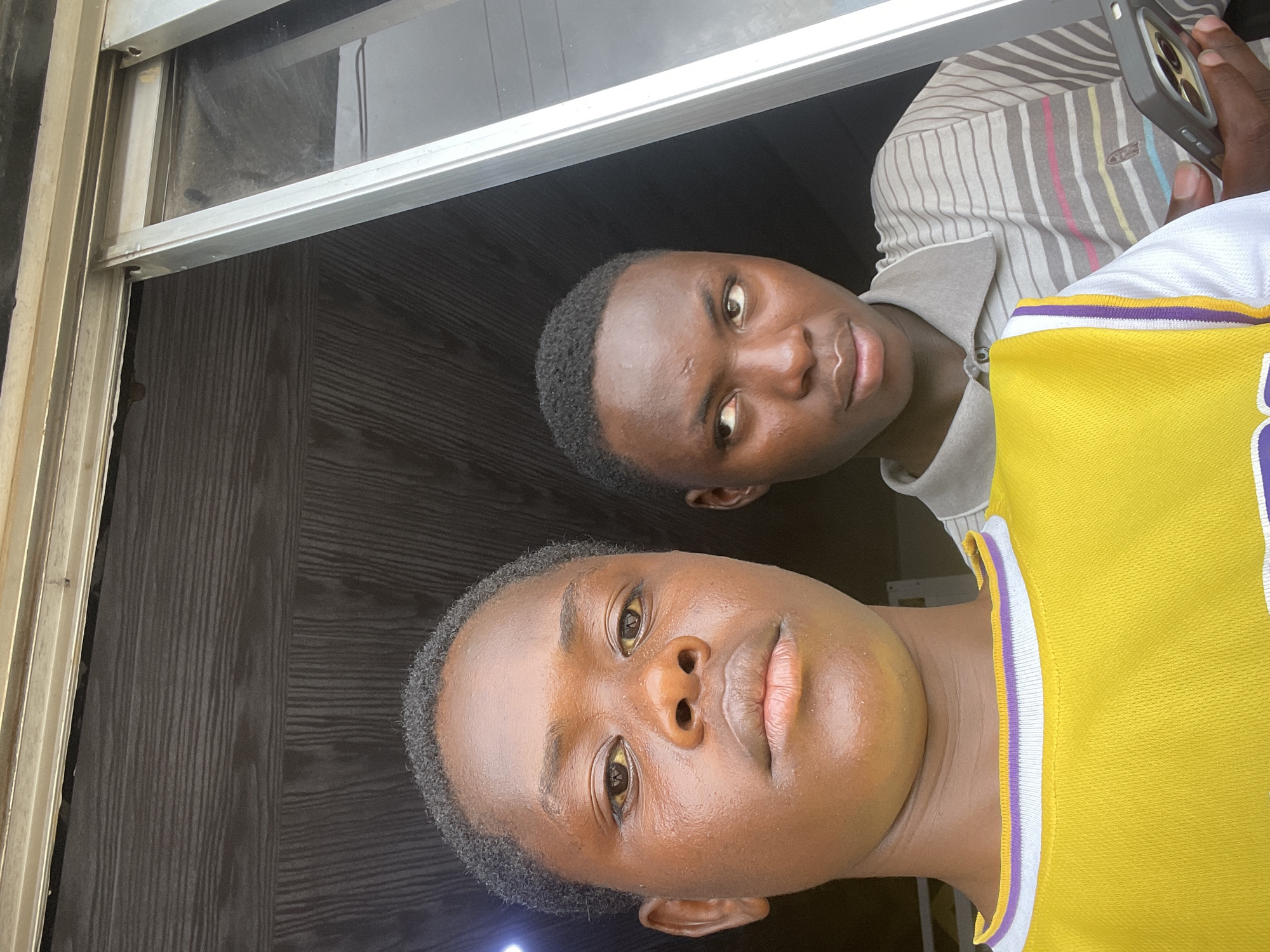 The image depicts two young Black males, likely teenagers, posing for a selfie. The foreground is dominated by the face of the individual on the left, who is looking directly at the camera. They have dark skin, short, dark hair, and are wearing a bright yellow t-shirt with purple and white trim around the neckline. The individual on the right is positioned slightly behind and to the right of the first. They have their head turned to the side, looking off-camera, and are wearing a light-colored polo shirt with thin horizontal stripes in various colors. This individual is holding a smartphone in their right hand.
The background consists of a wooden surface with horizontal grain, suggesting an indoor setting or a covered outdoor space. A silver-colored sliding window frame is visible on the right side of the image, separating the two individuals from an unseen exterior. The lighting suggests it is daytime, with some light coming from the right. The overall impression is of a casual, informal moment captured in a residential or commercial area in Jos, Nigeria.