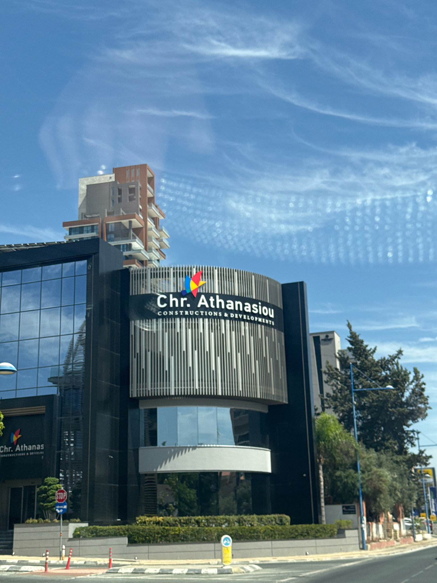 The image shows a modern office building with a dark facade and glass windows, featuring the company name "Chr. Athanasiou CONSTRUCTIONS & DEVELOPMENTS" prominently displayed. A colorful logo with red, yellow, and blue elements is also visible above the company name. The building is situated in a city environment, with a clear blue sky filled with wispy white clouds. Trees and other vegetation are present around the building, and a street with road markings and traffic signs is in the foreground. In the background, another taller building with a more intricate architectural design is visible. The scene appears to be an exterior shot of a commercial property.