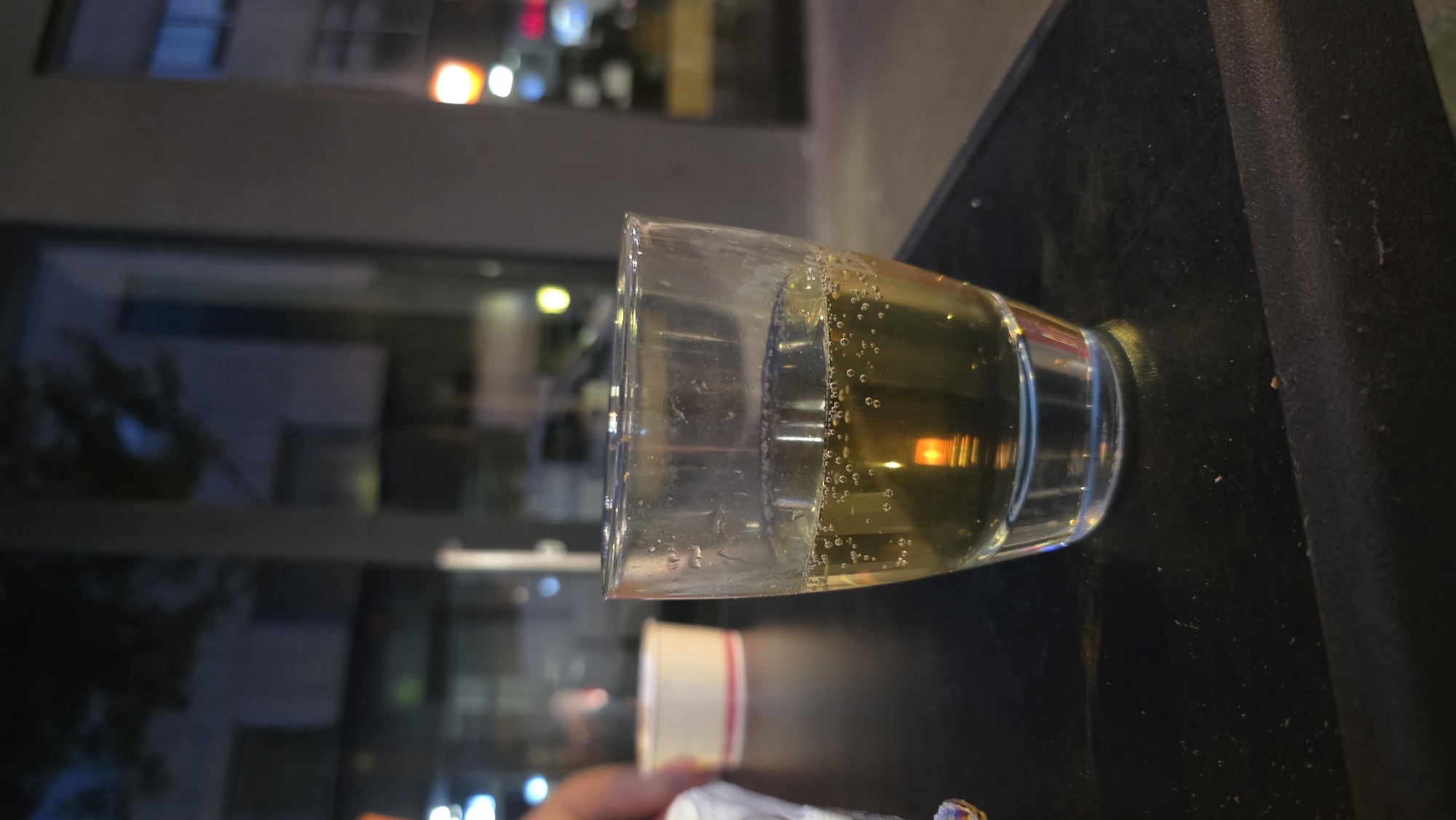 A close-up, eye-level shot shows a clear glass containing a light-colored alcoholic beverage, possibly a shot of liquor, with numerous small bubbles. The glass sits on a dark, textured surface, likely a table. In the background, a blurred scene depicts a building with windows reflecting lights, suggesting an urban nighttime setting. A white cup with a pink rim is also visible to the left of the glass, and a blurry hand is partially visible in the far left of the frame. The overall atmosphere is informal, possibly a bar or restaurant.