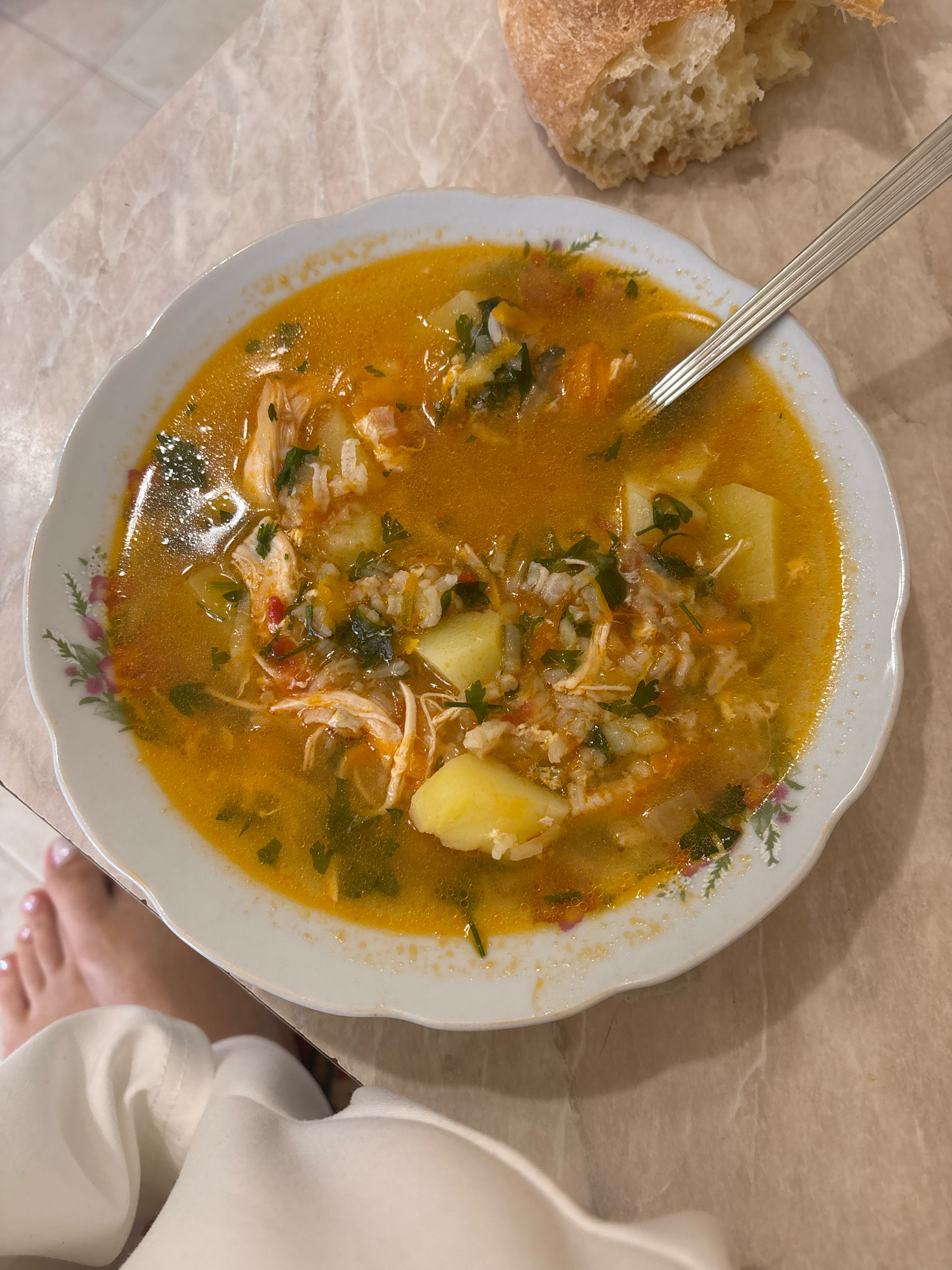 A close-up, overhead shot shows a white, floral-patterned bowl of chicken soup, filled with shredded chicken, rice, diced potatoes, carrots, and parsley, resting on a marble-patterned tabletop. A silver spoon is partially submerged in the soup. To the upper right of the bowl, a piece of torn crusty bread is visible. In the lower left of the frame, a person's bare foot with white-painted toenails is visible, next to a white cloth. The scene appears to be in a dining setting within a residence in Getahovit, Armenia.