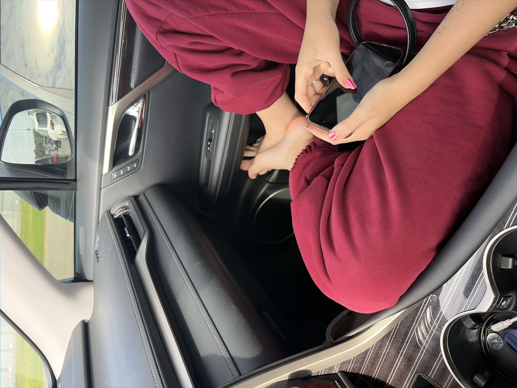 The image shows the interior of a car with a woman's bare feet resting on the floor in front of her. She is wearing a burgundy-colored outfit. Her hands are visible, holding a smartphone. The car's dashboard, door panel, and a side mirror reflecting the outside are also visible. The exterior view in the side mirror shows a road with other vehicles and a bright sky. The scene appears to be captured during daylight hours. This image does not provide any information about the city of Balashikha, Russia.
