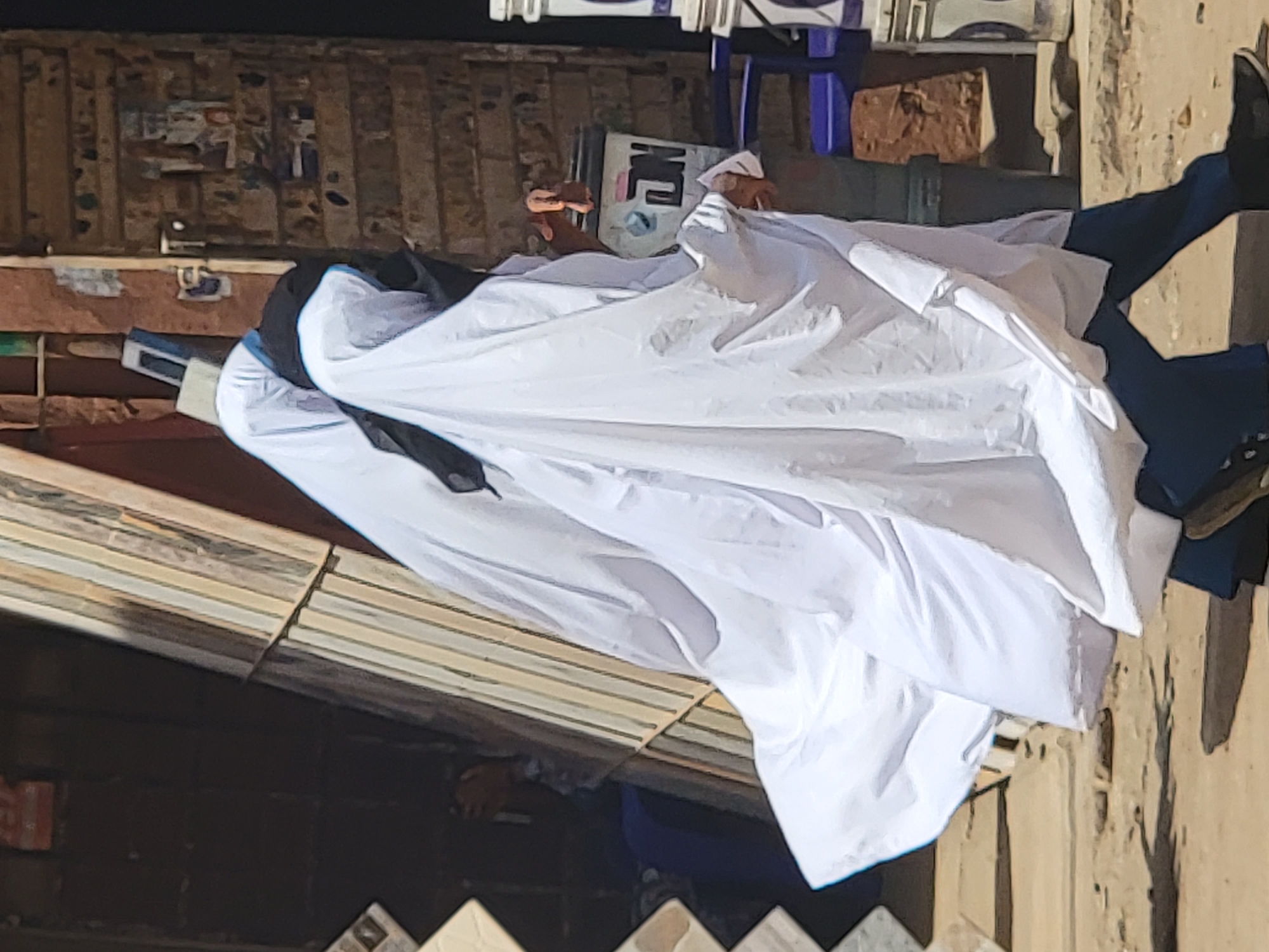 A person wearing a white garment and dark trousers stands outside a building in Maiburiji, Nigeria. The building exterior is a mix of brick and textured wall surfaces. The person seems to be interacting with the garment, perhaps adjusting or displaying it. Other items visible include a chair and various objects scattered around.