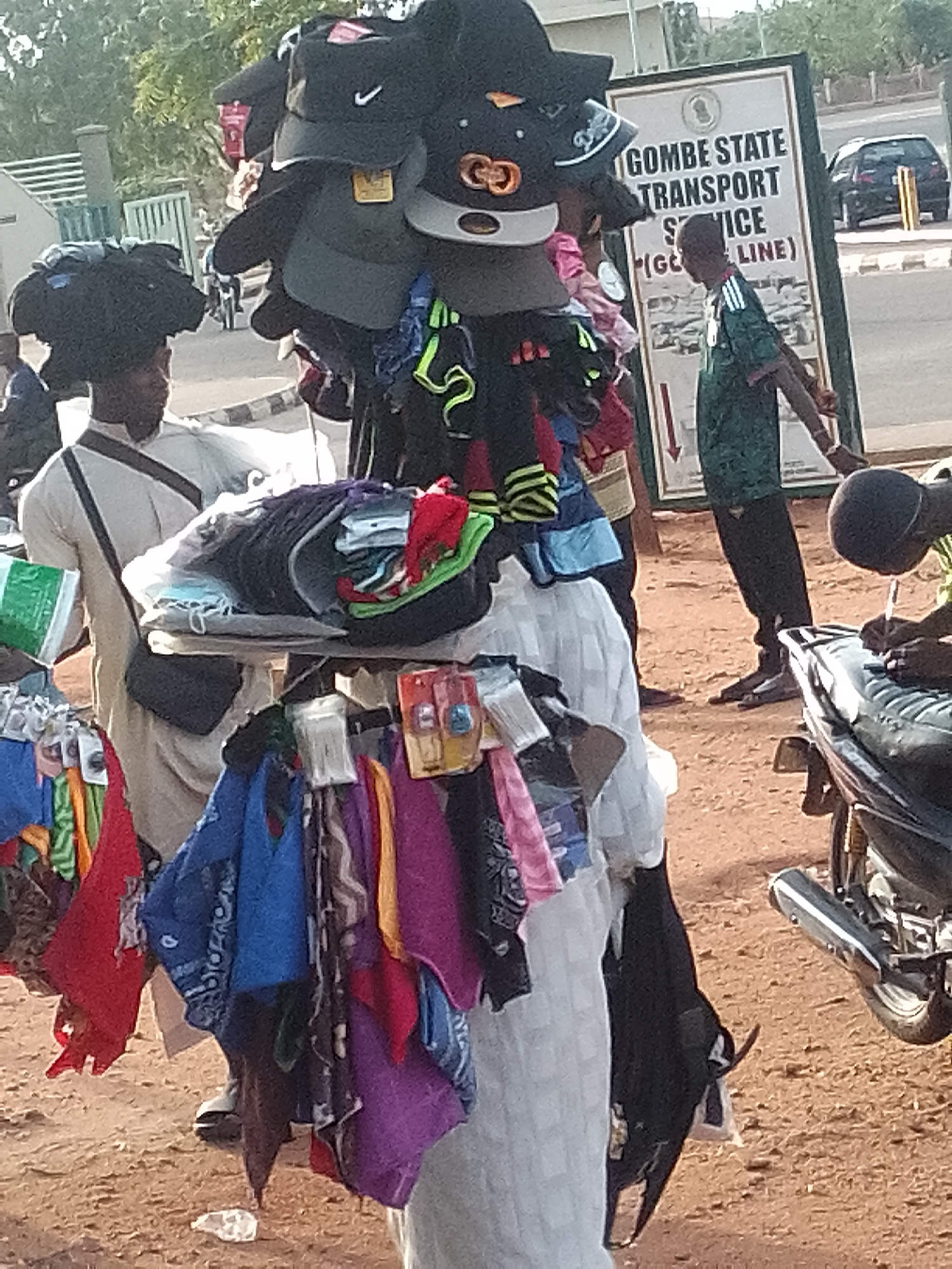 The image depicts a street scene in Gombe, Nigeria, likely near a transportation hub, given the "GOMBE STATE TRANSPORT SERVICE" sign. In the foreground, a vendor is displaying a variety of items, primarily hats and colorful textiles, arranged on a tiered rack. Several individuals are present. One person, wearing a light-colored robe and a dark bag across their chest, is standing behind the display of goods. Another person in a green and white striped shirt is standing near the sign. To the right, another individual, partially obscured, appears to be sitting on a motorcycle and is engaged in an action that resembles writing or inspecting something. The ground is covered with reddish-brown soil, characteristic of many parts of Nigeria. In the background, trees and a building with a fence are visible, along with a black car parked on the side of the road. The overall atmosphere suggests a busy, everyday commercial environment.