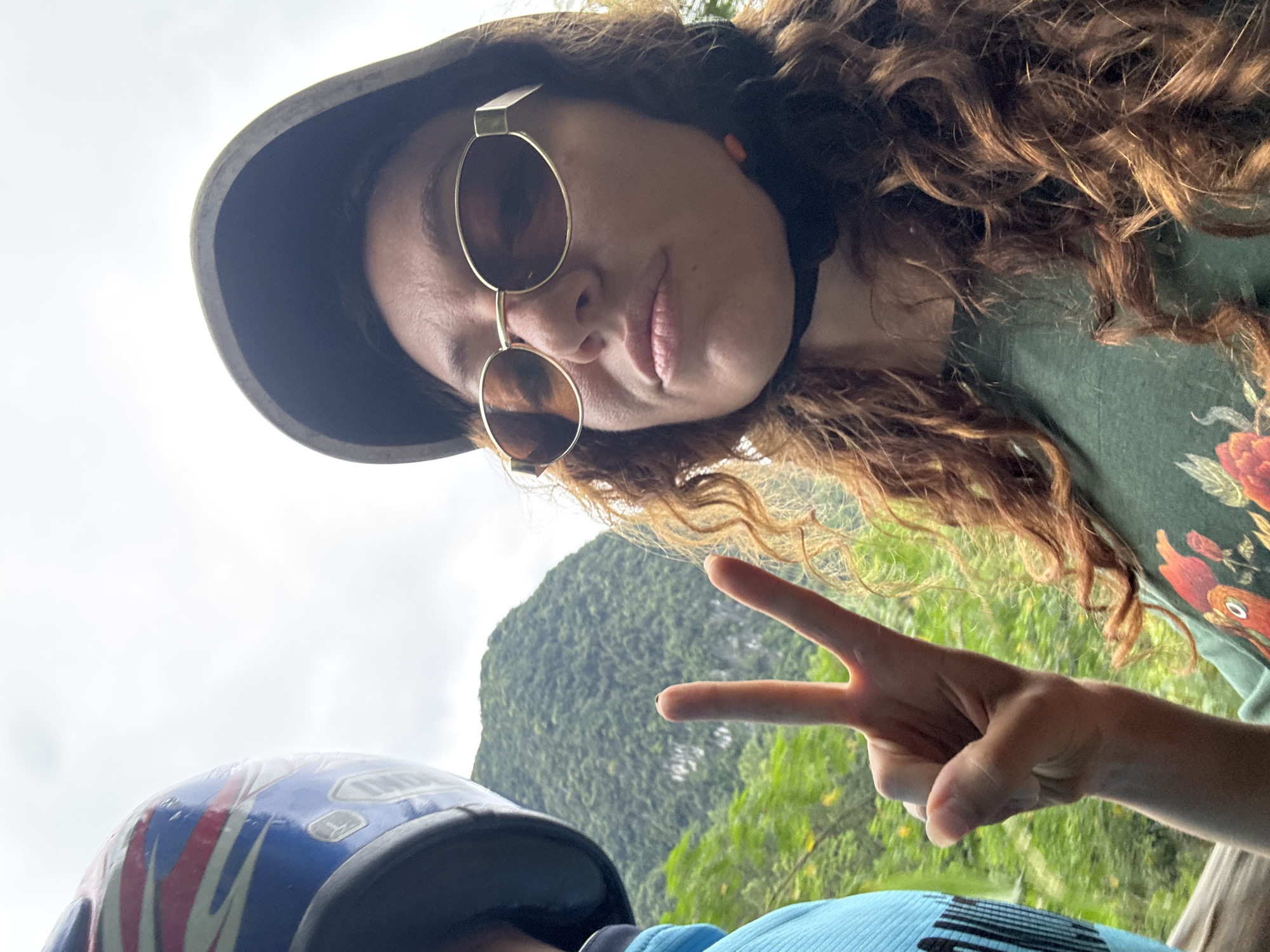 In an outdoor, overcast scene with a mountainous backdrop covered in dense green foliage, a person is taking a selfie. They are wearing a dark helmet and sunglasses with golden frames. The person's right hand is raised, fingers forming a peace sign. To the left, another person is partially visible, wearing a blue top and a blue, red, and white patterned helmet. The overall impression is that they are engaged in an outdoor activity, possibly on a motorbike, in a lush, natural environment.