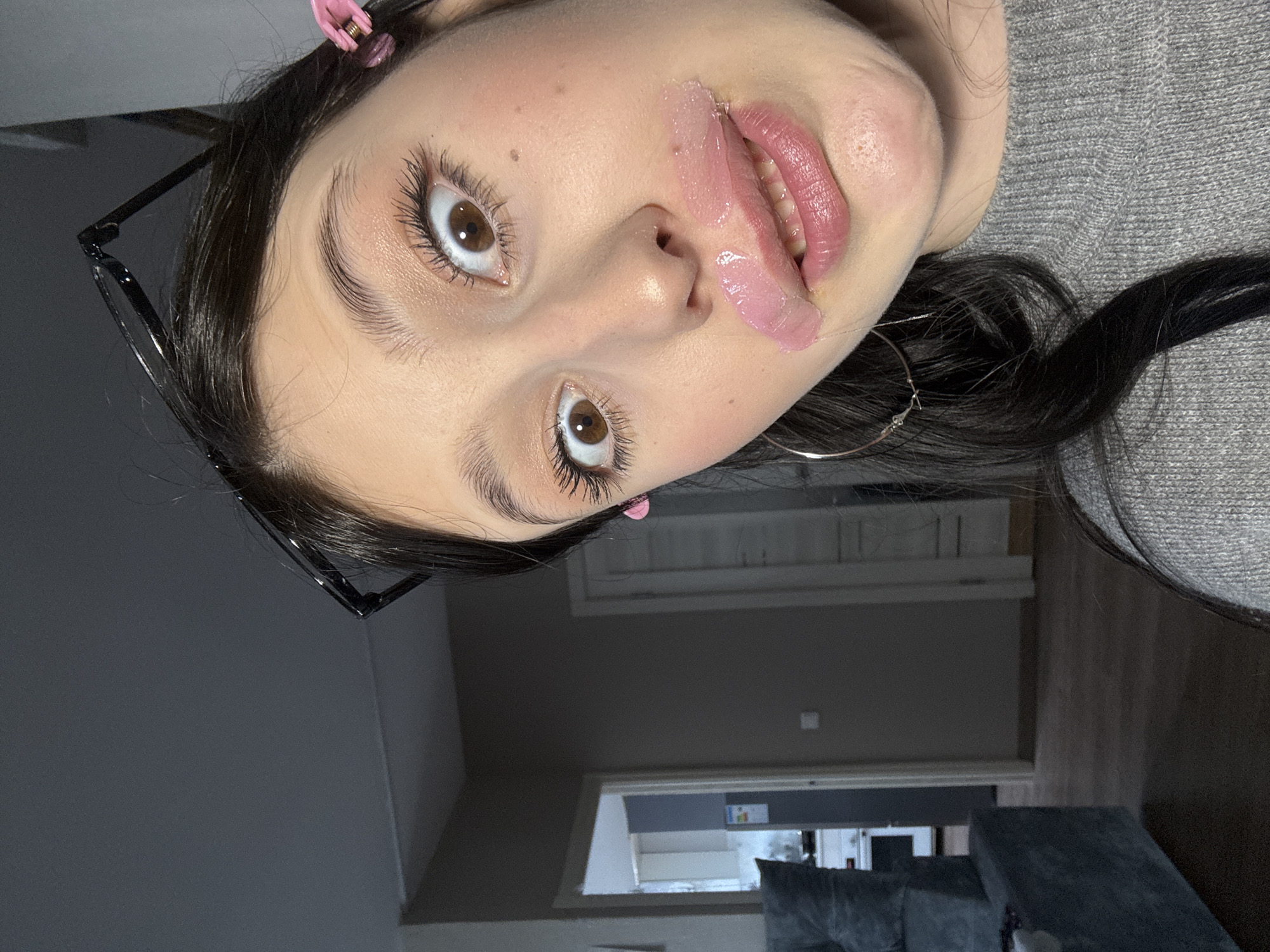 This image depicts a close-up, high-angle selfie of a young female subject taken indoors in a residential setting within the city of Chaikovsky, Russia. The subject has dark brown hair, dark brown eyes widened in a direct gaze, and a light complexion with subtle makeup on her eyelids and cheeks. A substantial amount of viscous, glossy, pink substance, consistent with a lip mask or heavy gloss, is applied to her lips, extending unevenly above the upper lip and appearing to drip or smudge downwards from the left corner of her mouth. Her lips are slightly parted, revealing some teeth. Black-framed eyeglasses are pushed up onto her forehead, and two small pink hair clips are visible in her hair above her right ear. A silver-colored hoop earring is visible on her left side. She wears a grey knitted sweater. The background features a dimly lit interior with dark grey walls and dark wooden or laminate flooring. A dark grey upholstered sofa is partially visible on the left. A doorway beyond the sofa leads into another room, likely a kitchen, identifiable by faintly visible white cabinetry and an appliance. Another closed white door is visible further to the right in the background.