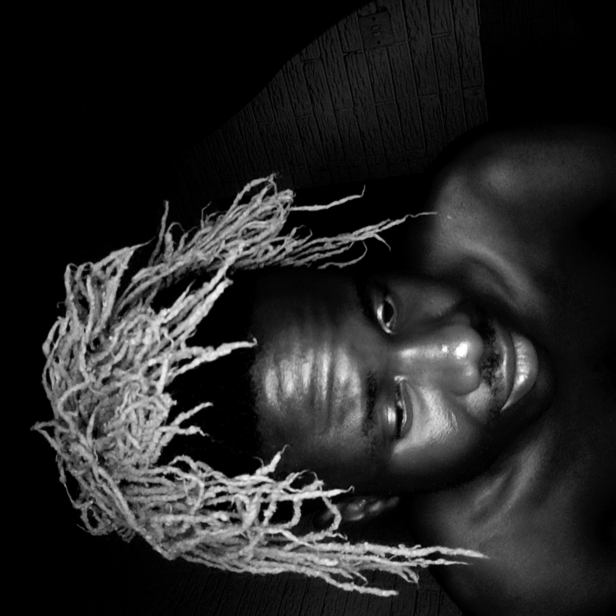 The image shows a close-up, low-angle black and white portrait of a person with light-colored dreadlocks. The individual is lying down, looking towards the camera. The background is dark, with a textured surface visible behind the head. The person's skin is glistening, and details of their facial features, including eyes, nose, and mouth, are clearly visible. There is no evidence to suggest a location in Narayi, Nigeria.