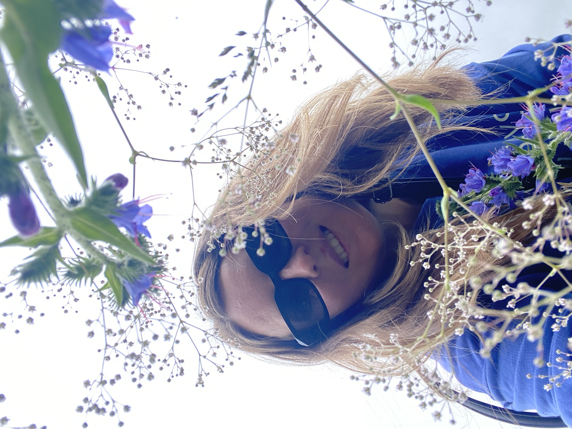 This is a close-up, low-angle shot of a woman with blonde hair and black sunglasses, wearing a blue sweatshirt. She is smiling and looking at the camera. She is surrounded by small white flowers and some larger purple flowers. The background is a bright, overcast sky. The scene appears to be outdoors, possibly in a park or garden, with no discernible city landmarks visible to confirm a location in Noviy, Russia.