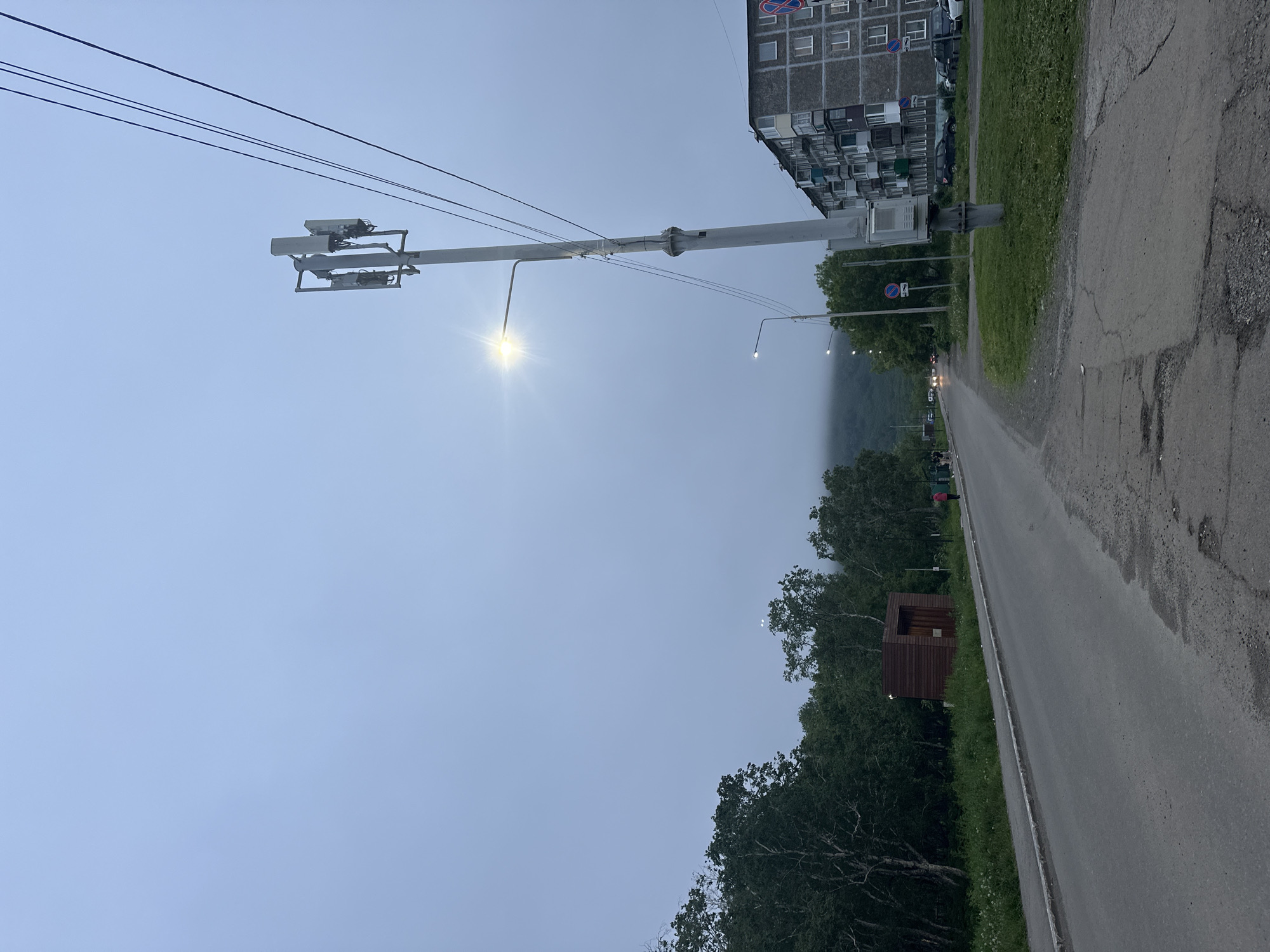 A street view captures a grey, overcast day in Vilyuchinsk, Russia. The foreground shows a cracked asphalt road with a sidewalk to the left. A line of trees borders the sidewalk, and behind them is a small, wooden, shed-like structure. Further down the road, a tall utility pole with a street light and cellular antennas is prominent. To the right of the pole, a multi-story apartment building with many windows and balconies is visible. Several cars are parked on the grass in front of the building. In the distance, a misty mountain looms. A few people are walking on the sidewalk and road, and cars are driving on the road. The overall atmosphere is subdued due to the cloudy weather.