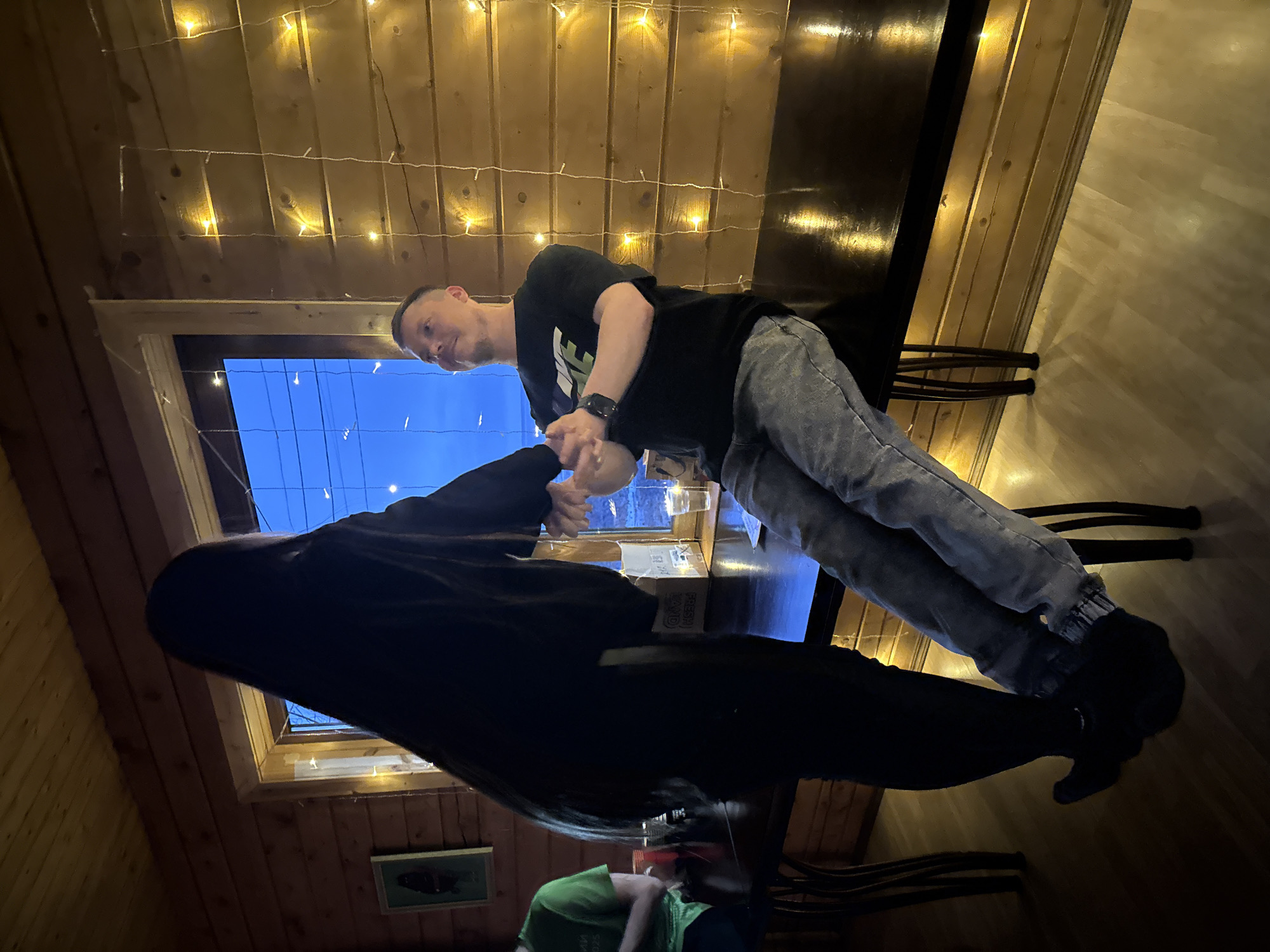 The image, taken in Chaikovsky, Russia, captures an indoor scene during the evening or night, indicated by the deep blue sky visible through a window. The setting features a warm, rustic ambiance with wooden paneling on the ceiling and walls, and string lights casting a soft, golden glow across the room.
In the foreground, a man wearing a black t-shirt with a white Nike logo, grey cuffed jeans, and dark shoes is leaning over a dark table or counter. He has short hair and a beard, and a gentle expression with a slight smile on his face. His left hand, adorned with a dark watch, is engaged with the hand of another person or an object on the table.
Opposite him, partially obscured by shadow, is another individual whose long, dark hair and dark clothing are visible. Their right hand appears to be interacting with the man's hand or a small, clear glass or cup on the table. A cardboard box with the word "FRESH" and other partially legible text like "KA O" or "KA VO" is also on the table.
In the lower left corner, a third person is partially visible, wearing a green t-shirt. Dark chairs or stools are glimpsed beneath the table. A framed picture, possibly depicting a fish, hangs on the wooden wall to the left of the scene. The overall impression is one of a casual, social gathering in a cozy, dimly lit environment.