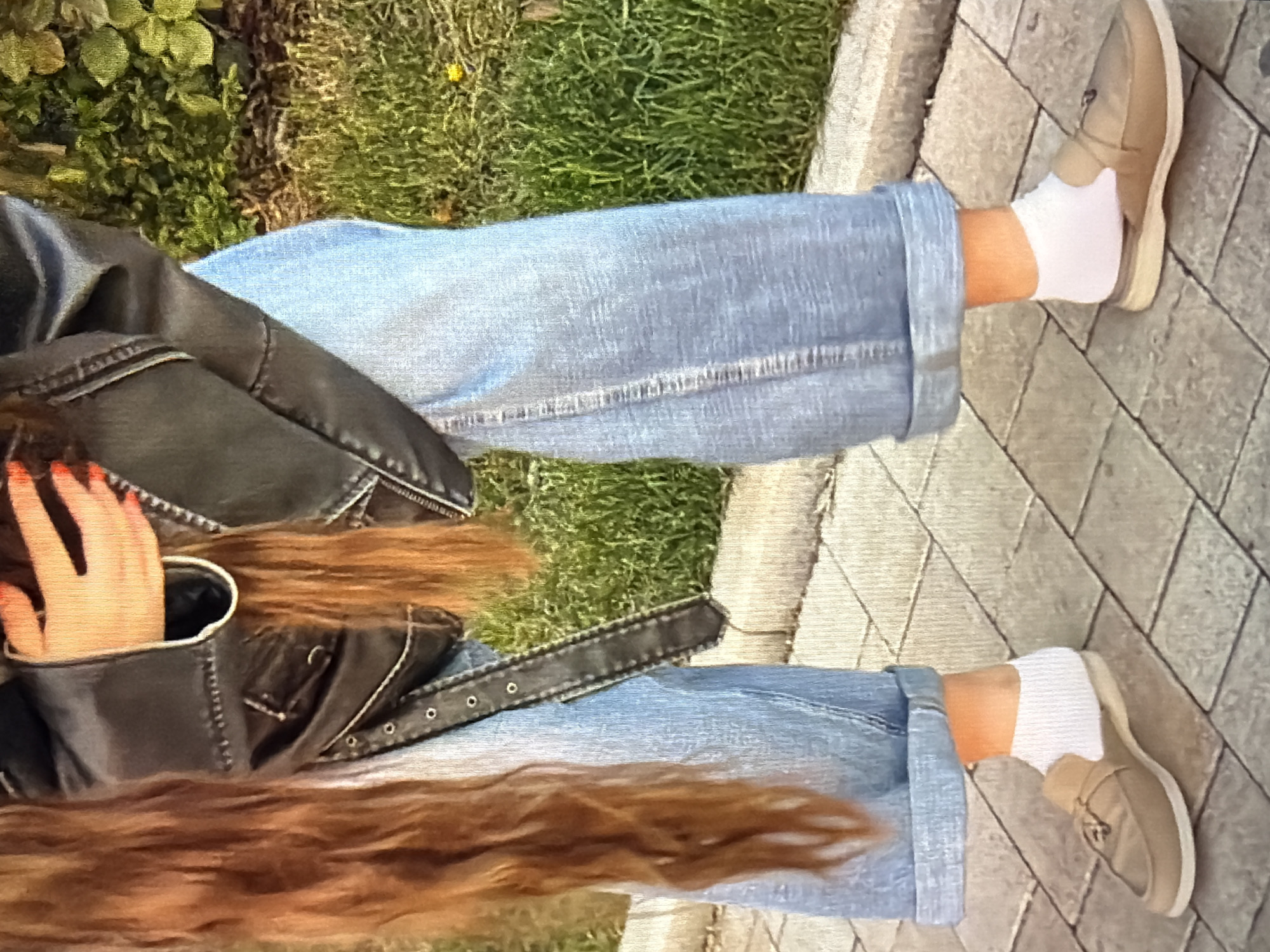 The image shows a person's legs and torso, seated outdoors. The person is wearing light blue, cuffed jeans, white socks, and tan slip-on shoes. They are also wearing a dark leather jacket. The person's long, reddish-brown hair is visible, along with their hand, which has bright red nail polish. The background consists of green grass and a paved walkway made of interlocking stones. The scene appears to be set during the daytime, with natural lighting. There is no visible text in the image.