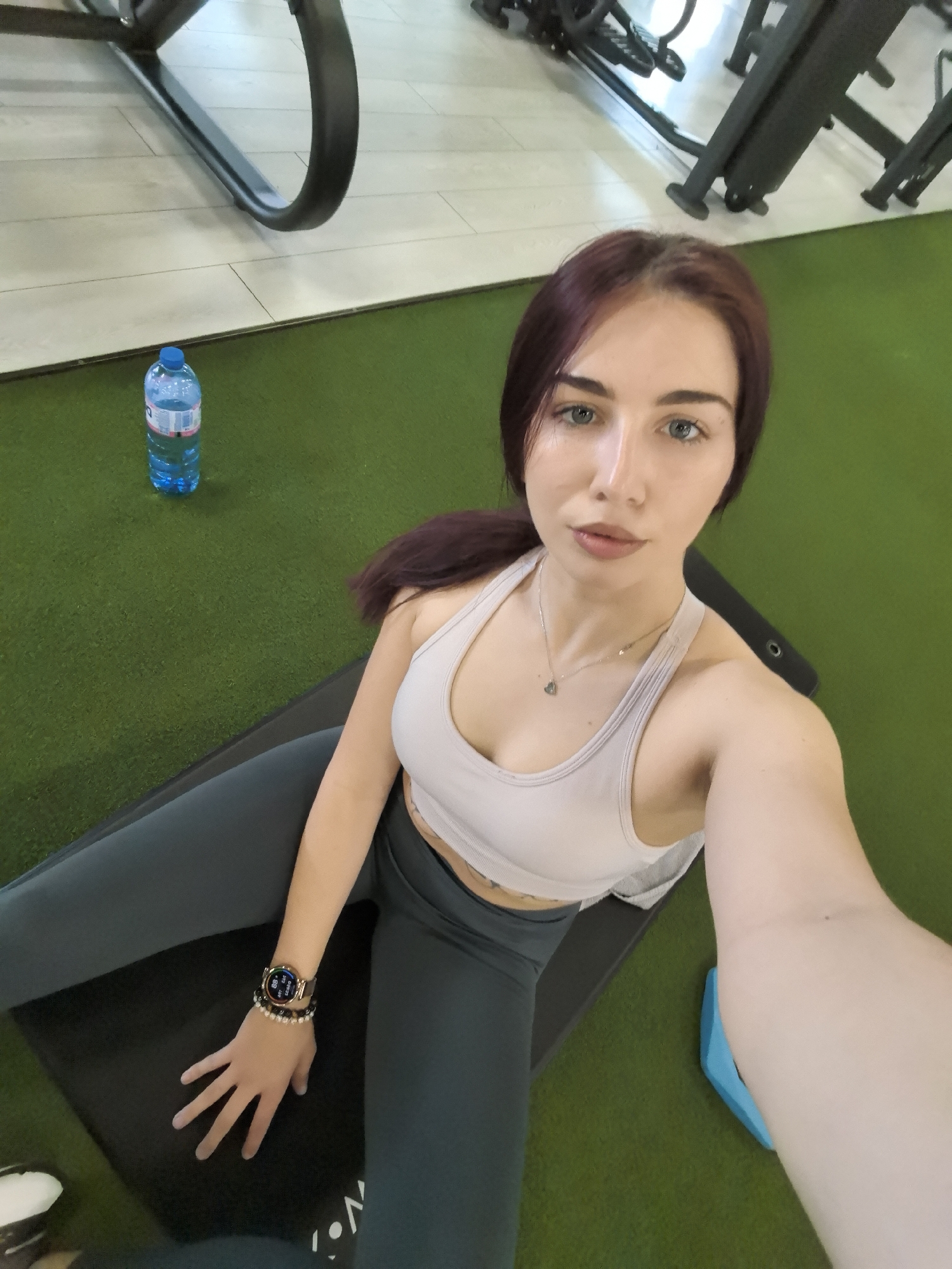The image shows a woman taking a selfie in what appears to be a gym. She is sitting on a black yoga mat, wearing a light-colored sports bra and dark leggings. She has reddish-brown hair pulled back in a ponytail, blue eyes, and a necklace. A smartwatch and beaded bracelet are visible on her left wrist. In the background, there's a bottle of water, a green floor, and gym equipment. The gym location is presumably in Stara Zagora, Bulgaria, though there is no specific element in the image that confirms this.