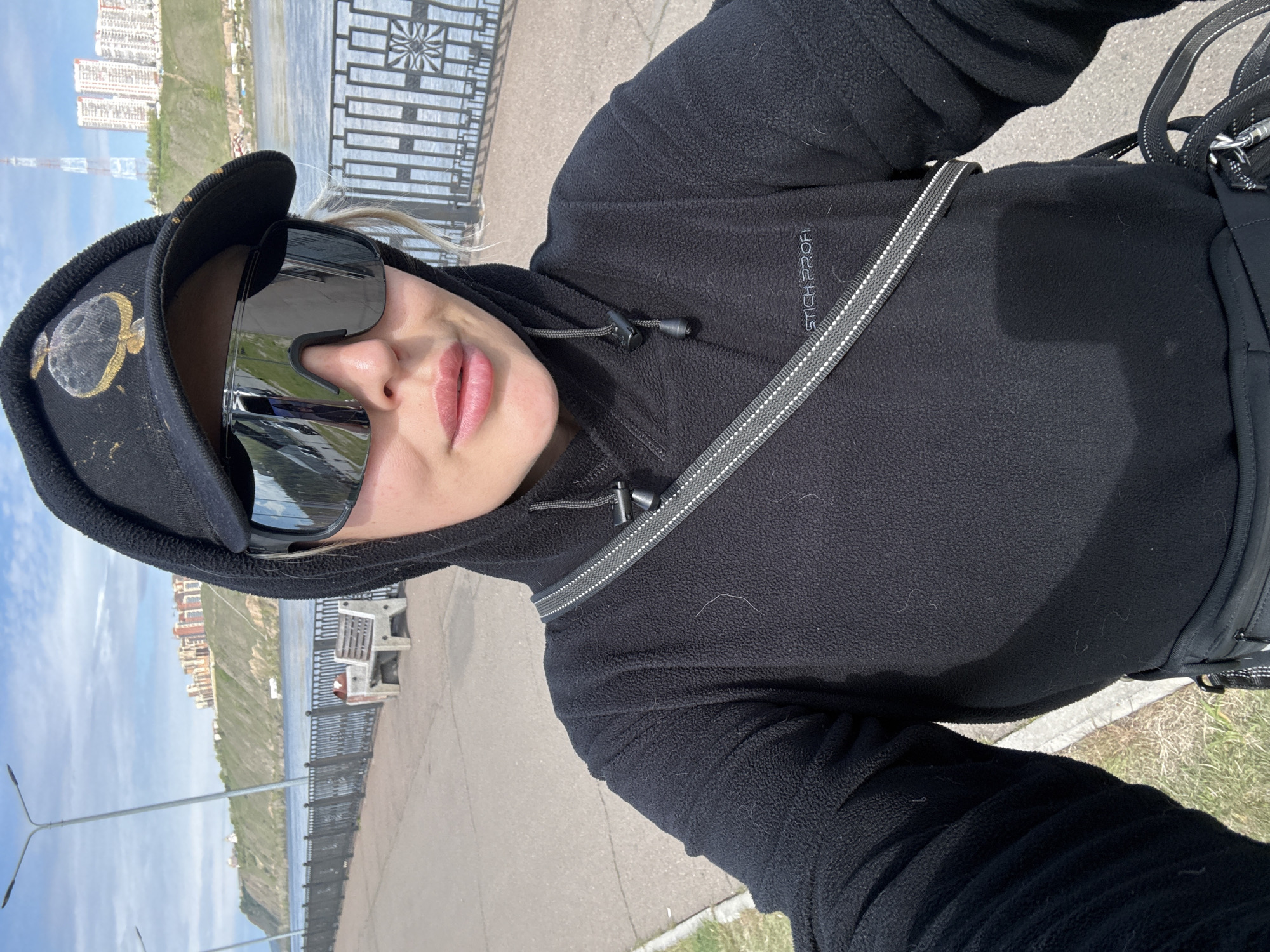 A selfie portrait shows a woman in a black hooded fleece jacket, black baseball cap with gold embroidery, and mirrored sunglasses. A grey and white strap crosses her chest. The background features an urban landscape with a river, a fence, and buildings. Specifically, this view is along the Yenisey River embankment, city of Krasnoyarsk, Russia.