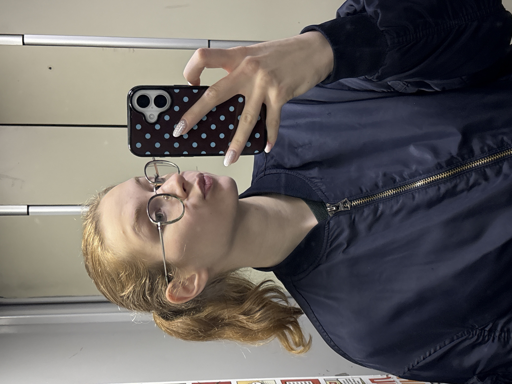 A young female with light brown hair tied in a ponytail is depicted taking a selfie. She wears wire-rimmed, round spectacles and a dark blue bomber jacket over a dark green garment. Her right hand holds a black smartphone encased in a dark cover featuring light blue polka dots; her index and middle fingers are extended over the phone screen, and her thumb is near the camera lens. Her fingernails are manicured with white, glitter-tipped extensions. Her lips are pursed in a "kissing" expression. The subject is positioned within an indoor environment characterized by light-colored, paneled walls, likely an elevator interior or a confined hallway. A vertical metallic or painted strip is visible on the wall behind her. On the far left, a vertical structure displaying multiple small, stacked red and white rectangular objects is partially visible. The precise location within the city of Belgrade, Serbia, is not identifiable from the visual evidence presented.