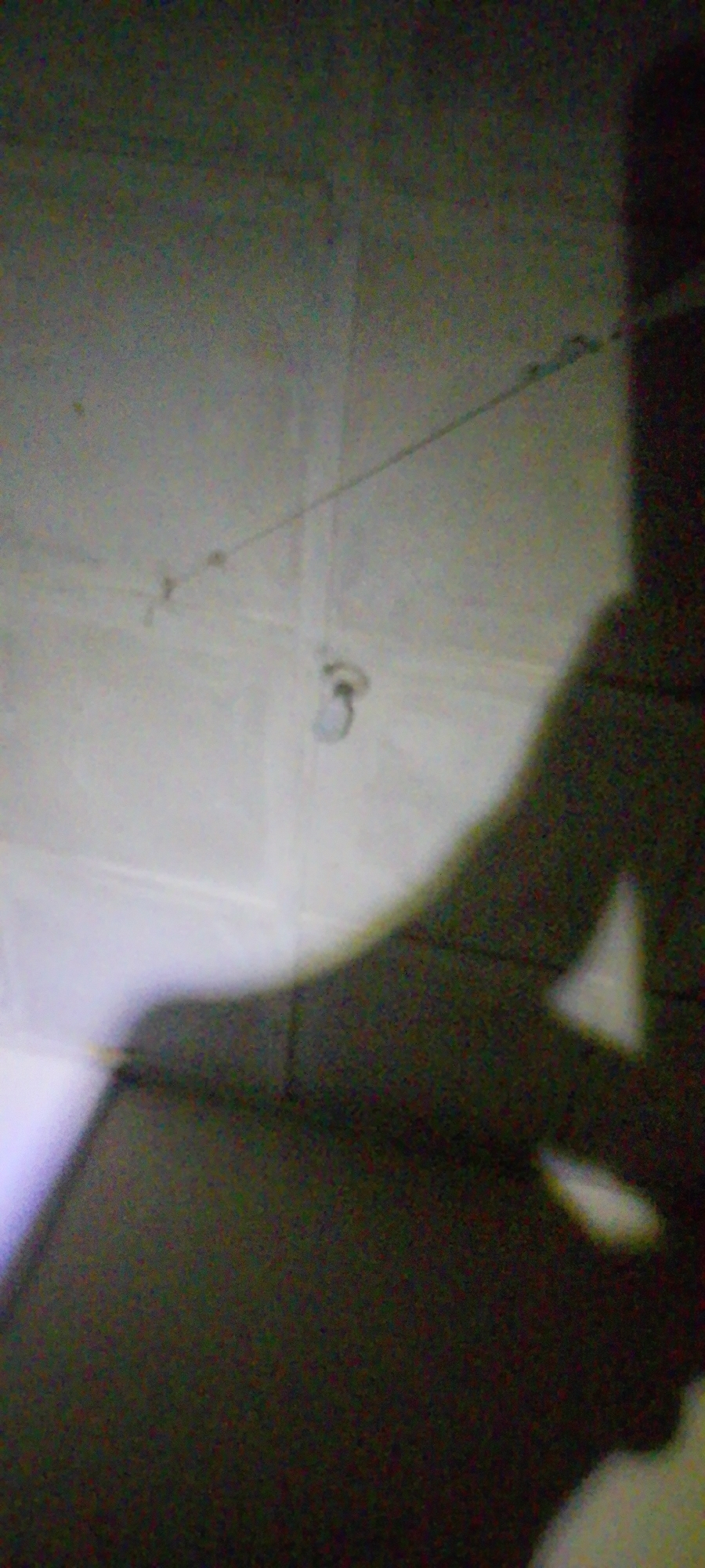 This is a blurry, low-light image of a ceiling with faint lines that suggest a suspended object or wires. A shadow is cast across the lower portion of the frame, indicating the presence of a person or object out of frame. The shadow's shape is indistinct but suggests an upright form. A small, light-colored object is visible hanging from a thin line near the center of the ceiling. The overall impression is that of an indoor space, possibly a room or corridor, with artificial lighting. The image quality is poor, with significant digital noise and blur, making it difficult to discern fine details. There is no visible text. The context of Jalingo, Nigeria, does not provide any specific visual cues within the image itself.