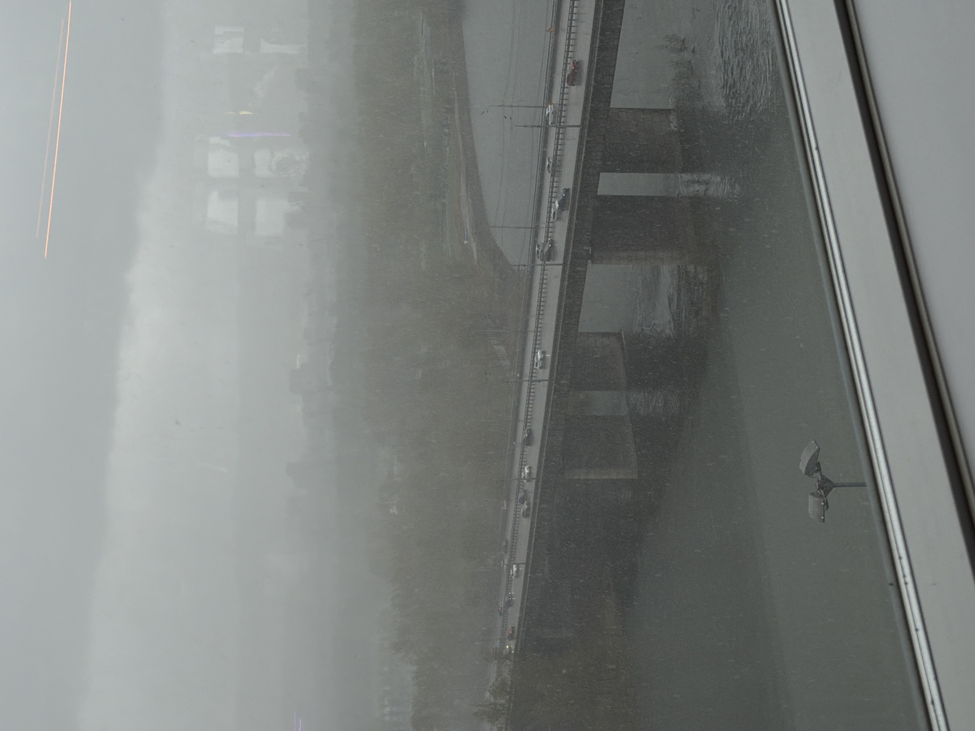 View from a window on a gray day. Looking out over a river with a bridge filled with traffic. The location is Krasnoyarsk, Russia. There is a city skyline visible through the fog. A weather station is visible on the right side of the image.