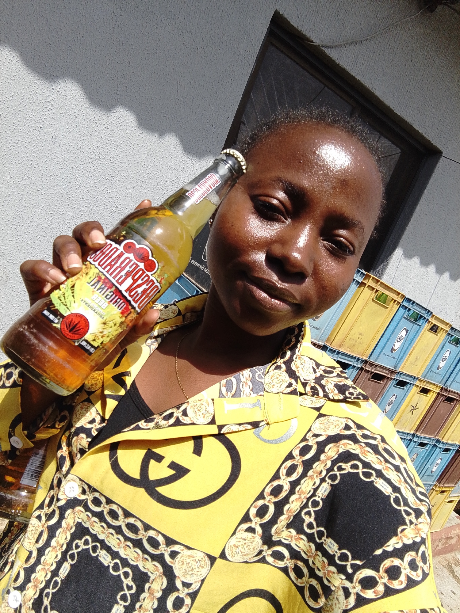 A young woman of African descent, with dark skin, short hair, and a light sheen of sweat on her forehead, is taking a selfie. She is holding a glass bottle of "Club Beer Original" in her right hand, tilted upwards and towards the camera. She is wearing a bright yellow shirt with a black and gold chain-link and crest pattern, and a black undershirt. The background shows a textured, light-colored wall with a shadow cast on it. To the right, a stack of yellow, blue, and brown plastic crates are visible. The scene appears to be outdoors, in what is likely a residential or commercial area, possibly in the city of Jos, Nigeria, given the specific brand of beer and typical stacking of crates. The lighting suggests it is daytime, with bright sunlight.