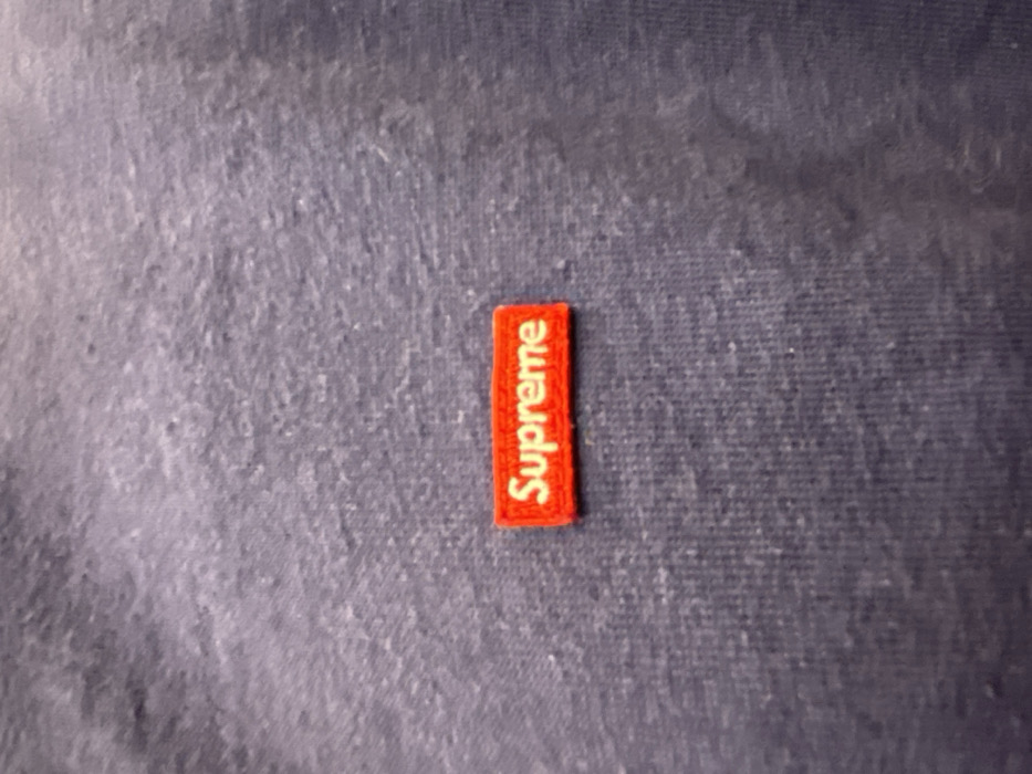A close-up image displays a small, red, rectangular brand tag embroidered with the word "Supreme" in white capital letters. The text is oriented vertically, reading upwards from the bottom of the tag. The tag is affixed to a textured fabric surface, which appears to be dark blue or grey, possibly a garment or textile item. The fabric exhibits some subtle variations in shading and surface texture, suggesting a worn or natural fiber. There are no people, discernible activities, or specific location cues within the image, beyond the fabric and the brand tag. The setting appears to be an indoor, neutral environment focusing solely on the object.