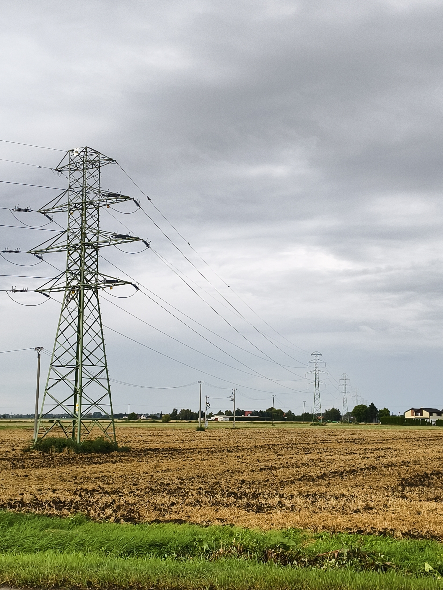The image depicts a wide, harvested field under a cloudy sky, with a series of electricity pylons stretching into the distance. In the foreground, a tall, green metal pylon stands prominently, with multiple wires extending from it across the frame. The field itself is covered in dry, brown stubble, indicative of a recent harvest. In the background, several more pylons are visible, diminishing in size as they recede into the landscape. A line of trees and some houses can be seen in the far distance, suggesting a rural or semi-rural setting. The sky is overcast with heavy, gray clouds. The image appears to be taken during the daytime. Based on external information, this scene is located in the vicinity of the city of Tczew, Poland.