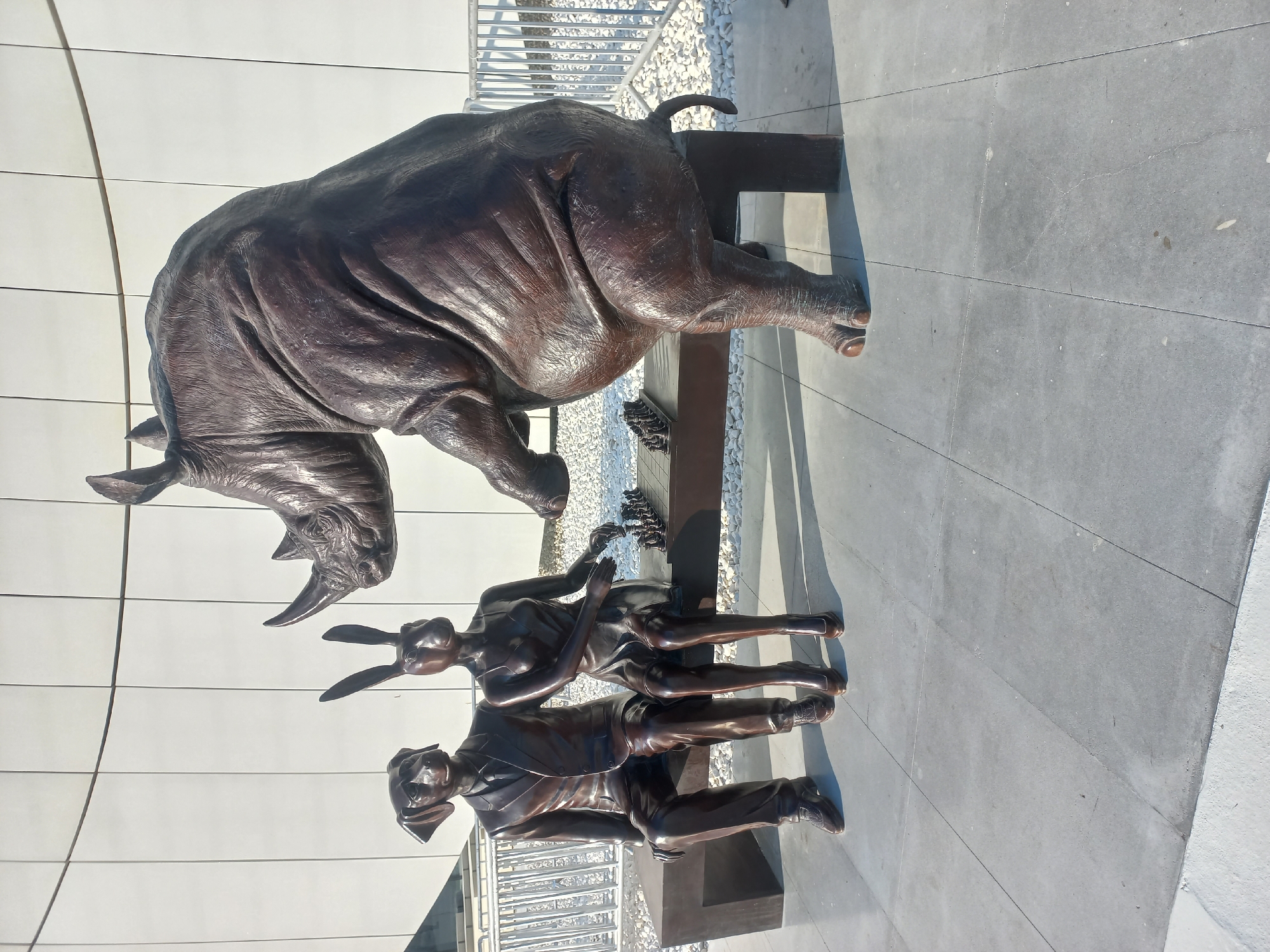 This is a photograph of a bronze sculpture located in the city of Baku, Azerbaijan. The sculpture depicts three anthropomorphic animal figures and a rhino playing chess. On the left, a dog-headed figure in a suit sits next to a rabbit-headed figure in a dress on a bench. To the right of the bench, a large rhino stands and appears to be making a move on the chessboard. The chess board is set up with pieces and placed on a bench. The background consists of a modern building with a white facade and some railings. The ground is paved with large, light gray tiles. The scene is illuminated by natural sunlight, casting shadows on the ground.