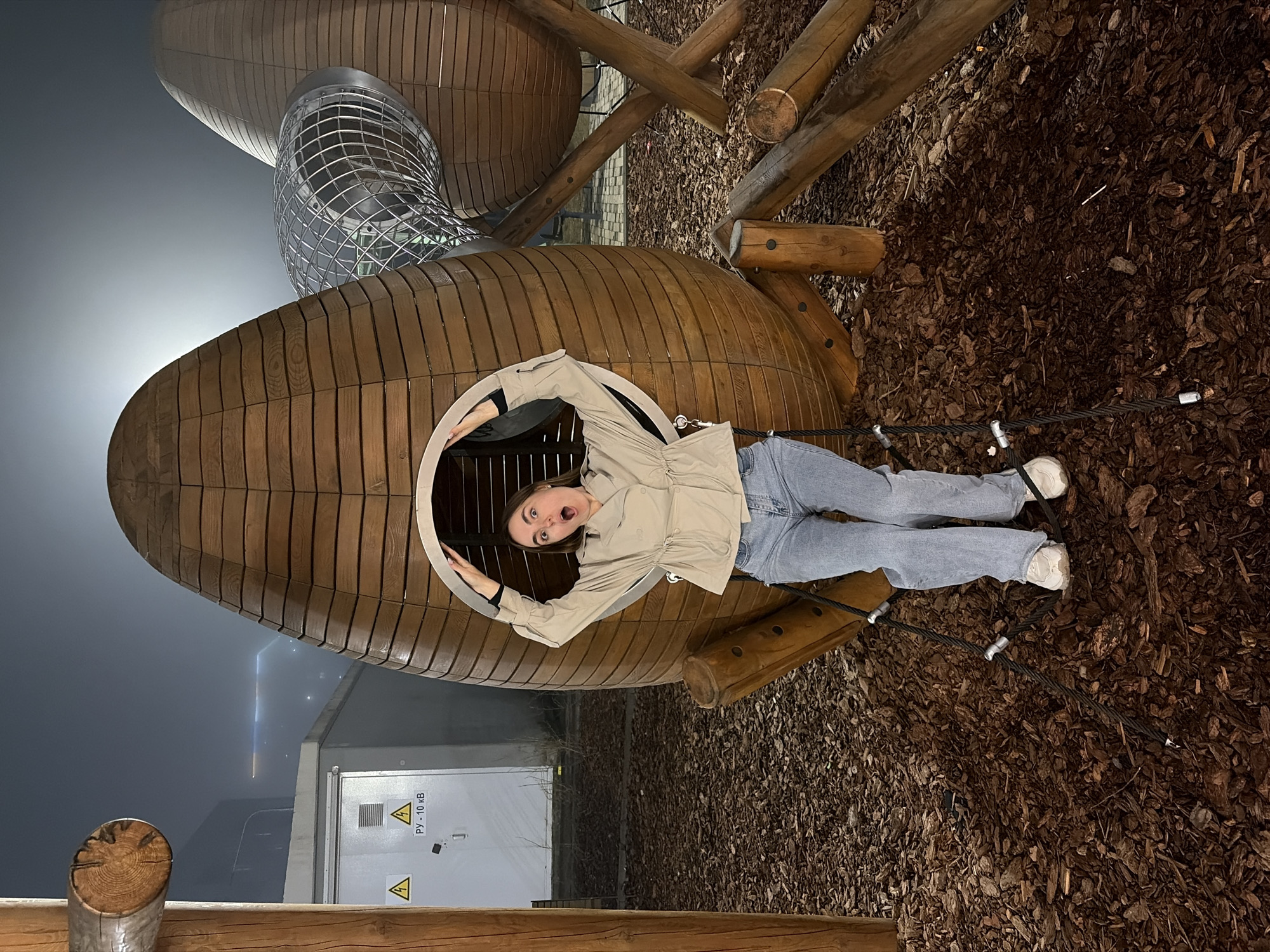 A person is inside a wooden structure in what appears to be a playground. The person, likely a female, has light skin and is wearing a beige jacket, blue jeans, and white sneakers. She is making a surprised or playful expression. The wooden structure is oval-shaped with a round opening, resembling a large pod or seed. A metal slide is connected to the wooden structure. The ground is covered with dark brown wood chips or mulch. In the background, there's a building with a power box labeled in Russian with voltage symbols, indicating a transformer. The surrounding environment has a foggy or dusky appearance, suggesting it's evening or night. The location is likely in Izhevsk, Russia.