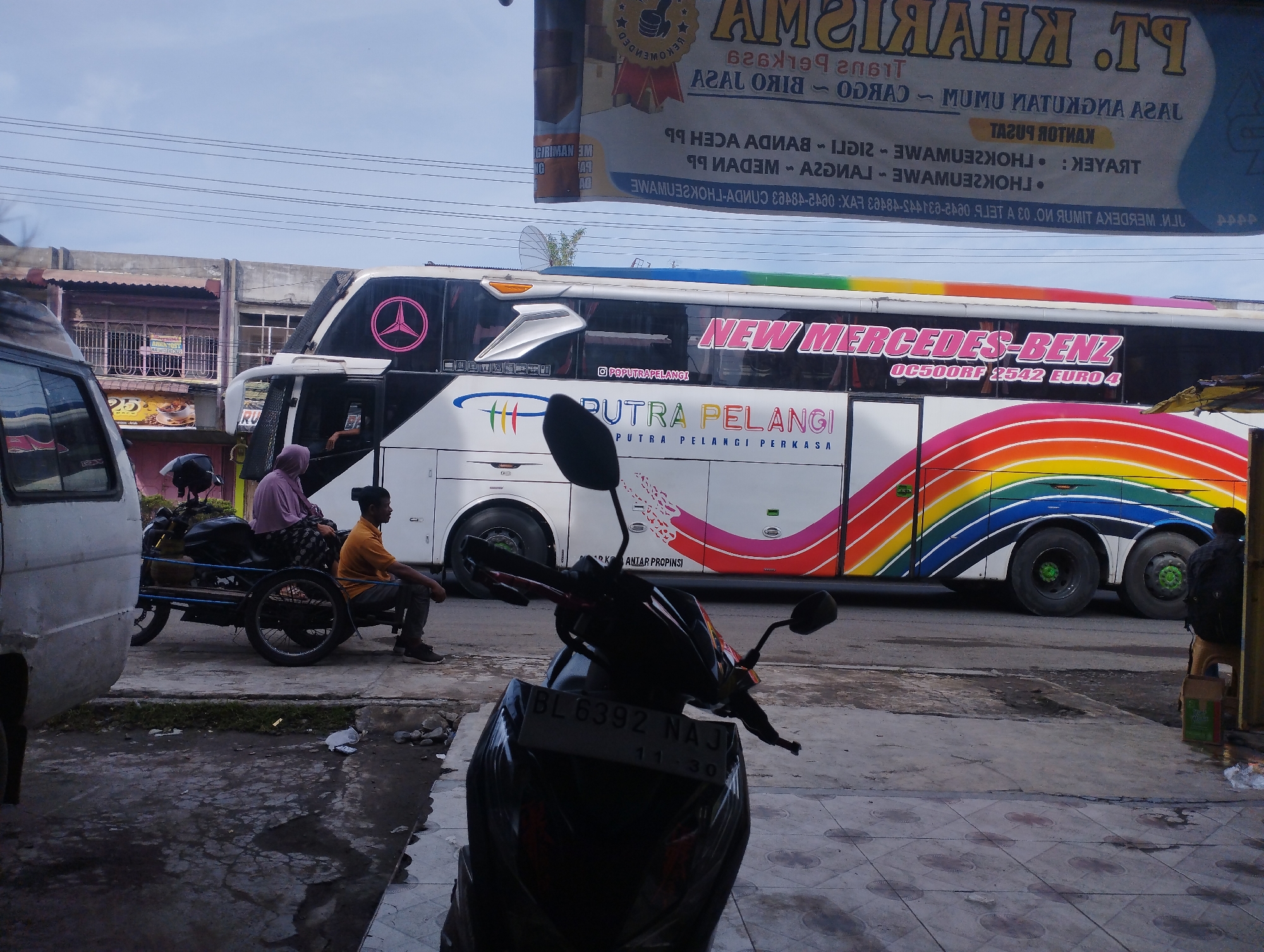 The image, likely taken in Lhokseumawe, Indonesia, shows a street scene featuring a "New Mercedes-Benz" bus adorned with rainbow stripes and the inscription "PUTRA PELANGI PERKASA." A sign overhead advertises "PT. KHARISMA," dealing in cargo and transportation. In the foreground, a parked motorcycle obscures some of the view. To the left, a van is partially visible. Further into the scene, two individuals are seated on a motorcycle-drawn cart. A man in an orange shirt is sitting on the cart while a woman dressed in a purple hijab also sits. There's also another man dressed in black, standing beside the bus. The background reveals shops and buildings, as well as power lines cutting across the sky.
