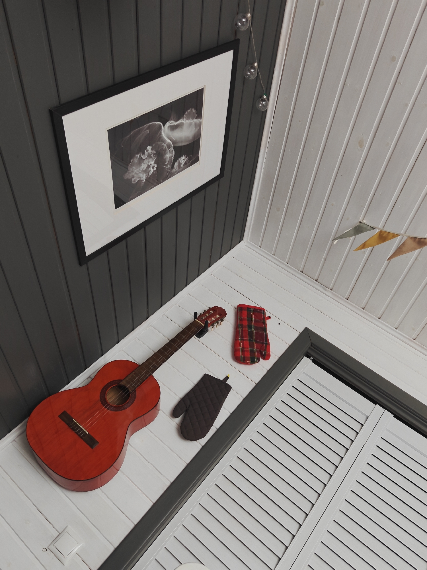 The image depicts an indoor scene with a dark gray paneled wall on the left and a white paneled wall and ceiling on the right. A framed black and white photograph of jellyfish hangs on the dark gray wall. In the foreground, a bright red acoustic guitar rests on a white wooden floor. To the right of the guitar are two oven mitts: one in a red and black plaid pattern and another in a dark quilted pattern. Further to the right, a louvered door or cabinet is partially visible. The white ceiling on the right has a string of small, clear light bulbs hanging down, and a few colorful fabric pennants are also visible near the ceiling. There are no people present in the image. The scene appears to be a domestic interior, possibly a room or hallway. The location of Koledino, Russia is not visually determinable from the image content.