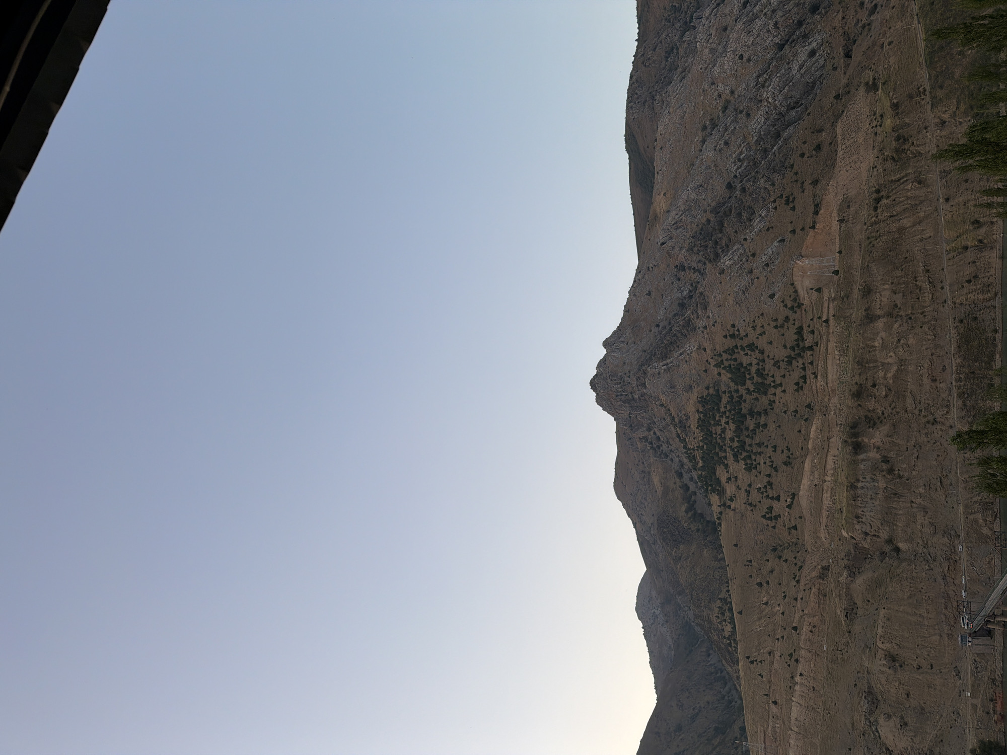 The image depicts a wide shot of a mountain range under a clear, pale blue sky. The mountains are rugged and dry, with sparse vegetation consisting of small, dark green trees scattered across their slopes. The terrain is characterized by a mixture of rocky outcrops and earthy, brown soil, with visible layers and erosion patterns.
In the lower part of the image, a body of water is visible, likely a river or lake. A bridge crosses the water, with a few vehicles, including what appear to be buses and cars, on it. A structure resembling a small dam or control tower is situated near the bridge. A few trees line the bank of the water body. A tall electrical pylon can be seen on one of the mountain slopes.
The overall impression is one of a natural, somewhat desolate landscape with signs of human infrastructure. The location is identified as Nanay, Uzbekistan. The lighting suggests it might be late afternoon or early morning due to the soft, even illumination and the clear sky. There are no people clearly discernible in the scene.