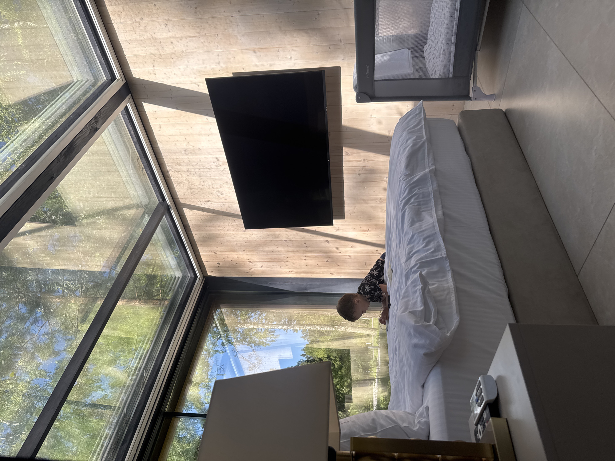 A child is playing on a bed in a room with a large window overlooking trees. The bed is white and located next to a crib. A television is mounted on a wooden wall above the bed. A lamp is in the foreground, partially obscuring the view of the window. The location is Ust'-Muny, Russia.