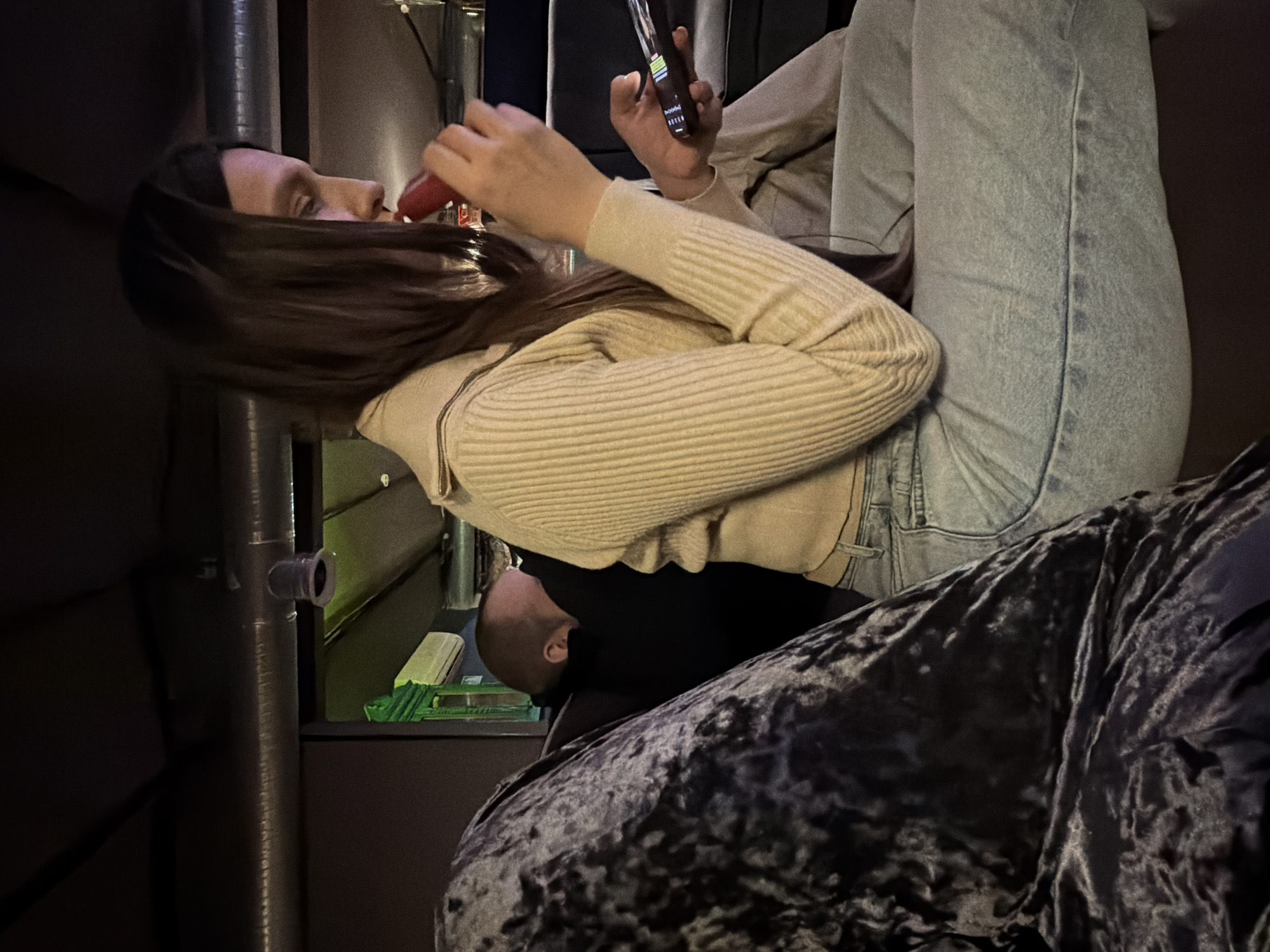 A woman with long brown hair sits on a dark patterned surface and looks at her phone. She wears a light-colored, ribbed sweater and light-wash jeans. In her other hand, she holds a small red object to her mouth. A bald man's head is partially visible below her. A metal pole is to the left, and a dark background is visible behind it. The location could be Moscow, Russia, but that cannot be confirmed from the image alone.