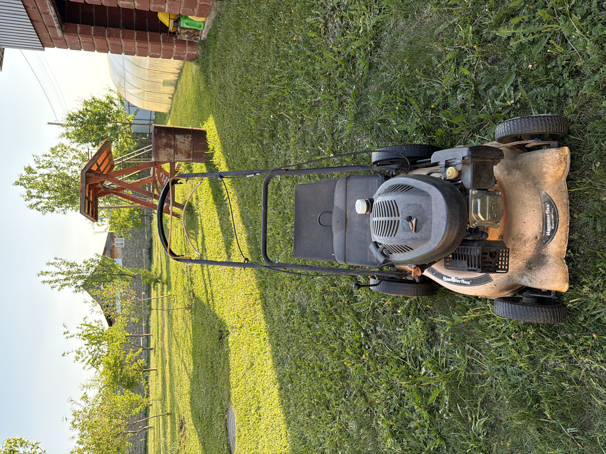 The image shows a lawnmower on a grassy lawn. In the background, there's a brick building, a metal frame structure, possibly for supporting plants, and what appears to be a greenhouse. In the far distance, there's a stone building with a window. The image was likely taken in a backyard or garden setting. There's no clear evidence to confirm the location is specifically Bolsheustyikinskoye, Russia, but the style of buildings is consistent with a rural area. The focus of the image is the lawnmower positioned in the center. The environment is primarily grassy, with some weeds interspersed. The lighting suggests it is daytime. There are no people visible in the image.