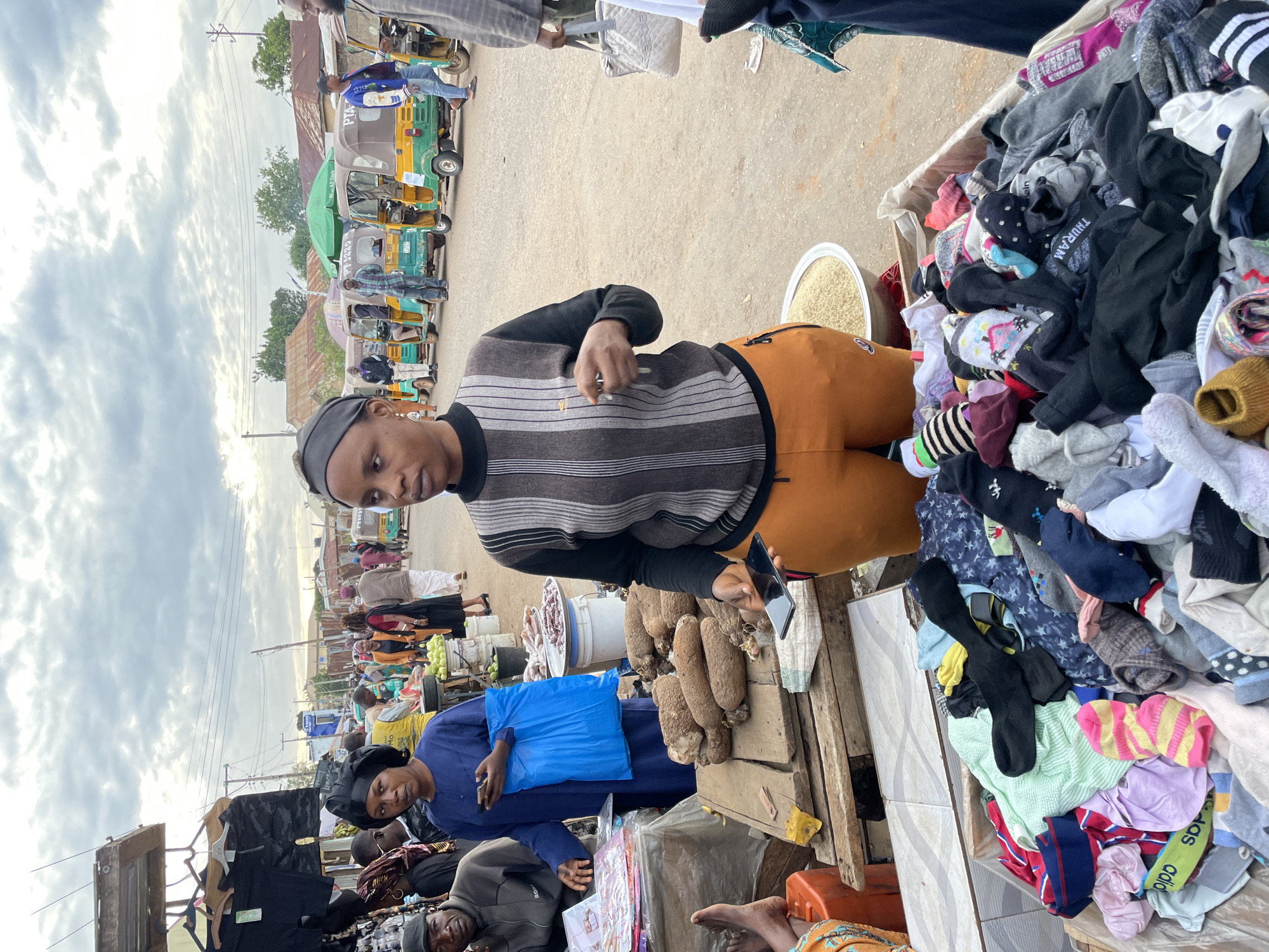 The image depicts an active outdoor market scene in Gura Top, Nigeria, under an overcast sky. In the foreground, a woman wearing a black and grey striped long-sleeved top and orange athletic pants, with a black head covering, stands facing slightly left, holding a dark smartphone in her right hand and a small reflective object in her left. Immediately in front of her is a large wooden crate filled with assorted socks and small textile items for sale. To her left, another woman in a dark blue gown and black headwrap stands behind a wooden stall displaying large brown root vegetables, likely yams. She holds a bright blue plastic bag and a smartphone. Partially visible to her left, a man in a dark jacket and cap is seated behind another stall with papers or magazines. His bare left foot rests on an orange container. A large metal bowl filled with light-colored grains is on the ground to the right of the central woman. In the midground, numerous individuals are walking and congregating amidst market activities. Several yellow and green auto-rickshaws ("Keke Napep") are lined up on an unpaved road in the background. Buildings with corrugated iron roofs line the street, and utility poles with overhead wires are visible.