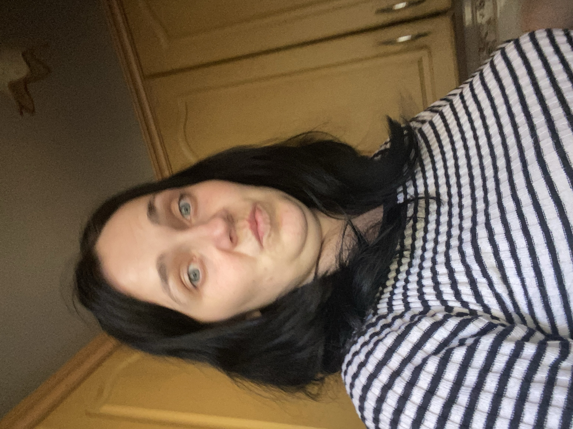 A woman with black hair and blue eyes is shown in a close-up shot. She is wearing a black and white striped shirt. Her facial expression is a pout, with her lips pursed. The background consists of what appears to be kitchen cabinets, painted a light yellow-brown. The lighting suggests an indoor setting, possibly illuminated by natural light from a window.